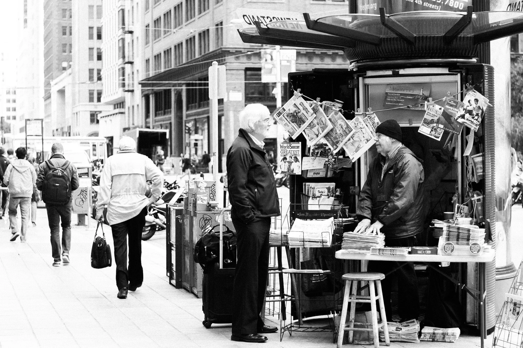 "Newsstand Conversation"
In a city of fast scrolls and silent feeds, a newsstand becomes a place of pause. Two men trade words beside headlines and old magazines—an exchange that feels timeless amid the blur of Market Street. This image honors the qu