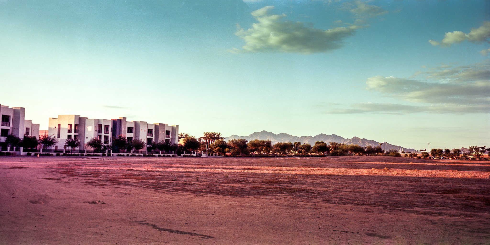 Where the Light Breaks: A band of morning light cuts across the empty lot, tracing the last open path to the mountain. The new development looms like a gate, neat and closed, while Estrella Mountain lingers beyond—distant, unchanged, and slowly edged