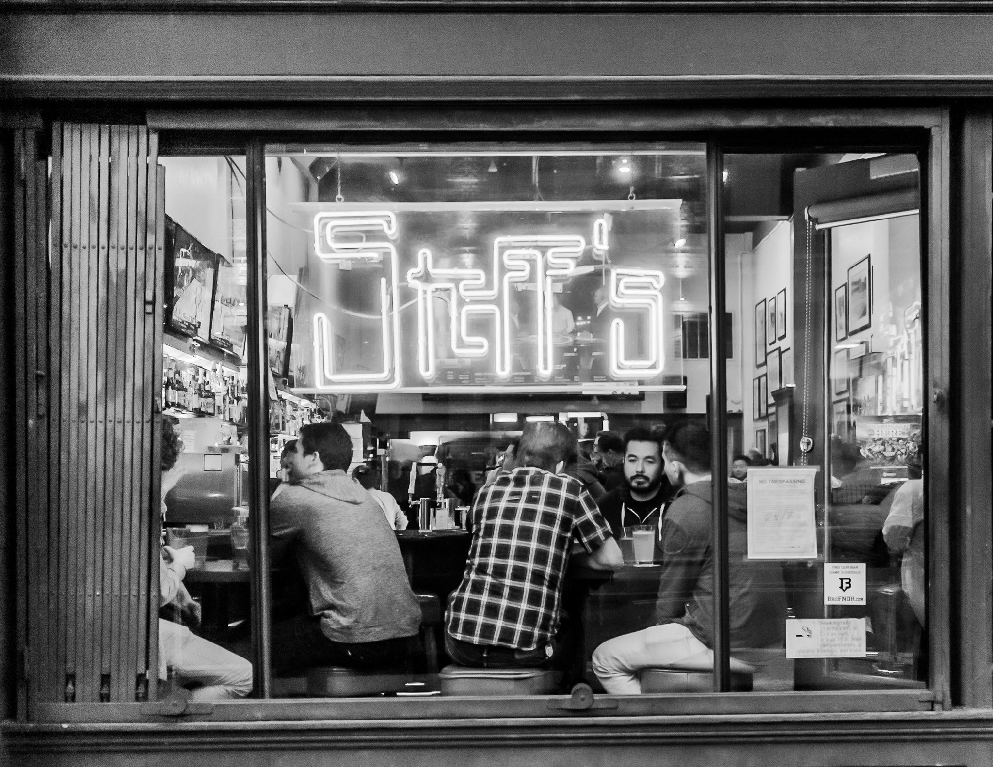 "Neon Conversations" Framed by a neon sign, a small group leans into the night—talking, waiting, drinking in silence. The moment feels both intimate and public, shared behind glass but open to the street. In a city known for movement, this is a quiet