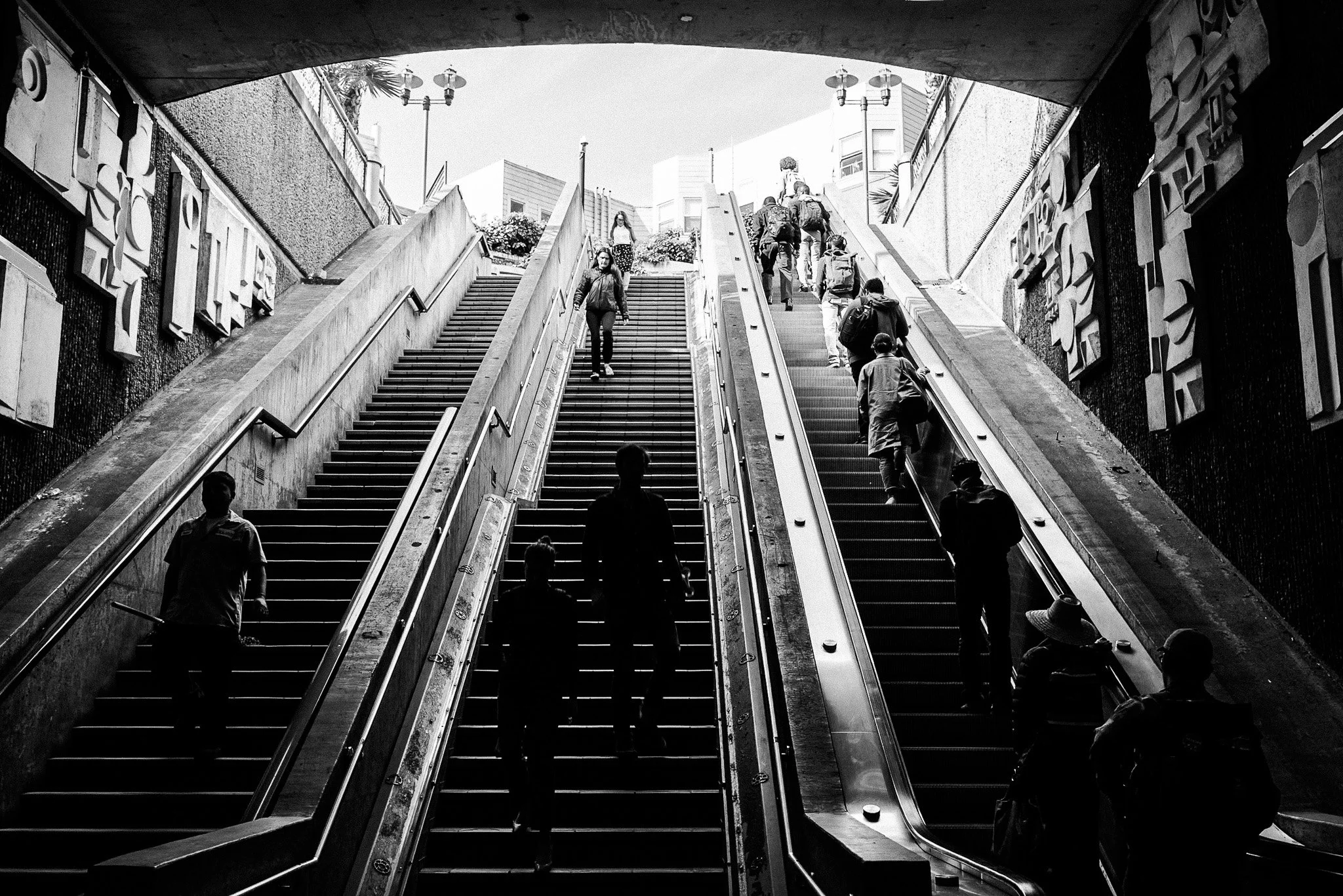 "Exit Above" Figures rise from shadow into daylight, scattered across stairs and escalator like notes on a scale. This moment captures that daily climb, where each person carries their own weight into the light.