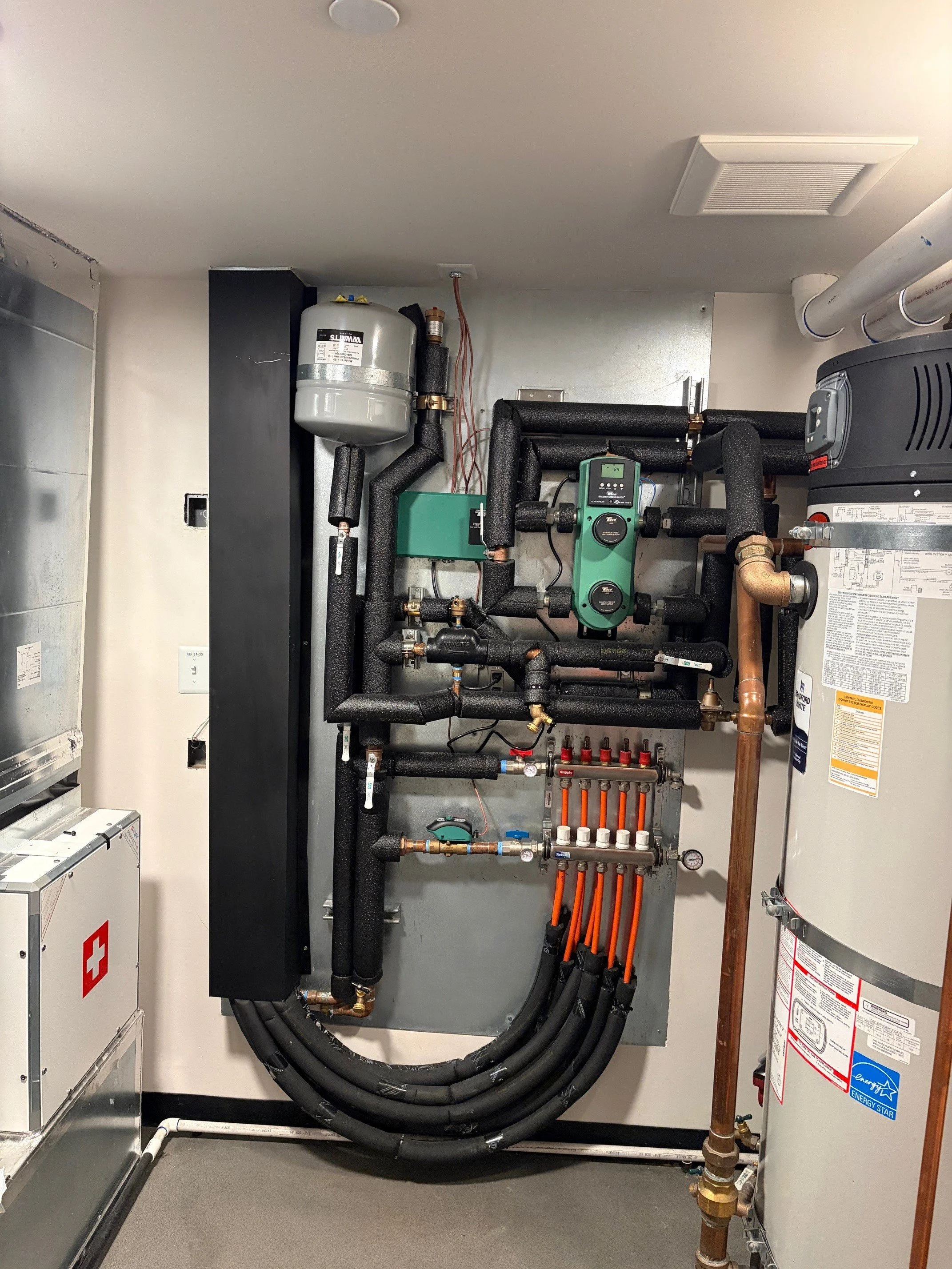 A mechanical room with a tank water heater, a circuit panel, a green HVAC control unit, and insulated black hydronic pipes with orange humidity tubing and copper hydronic pipes.