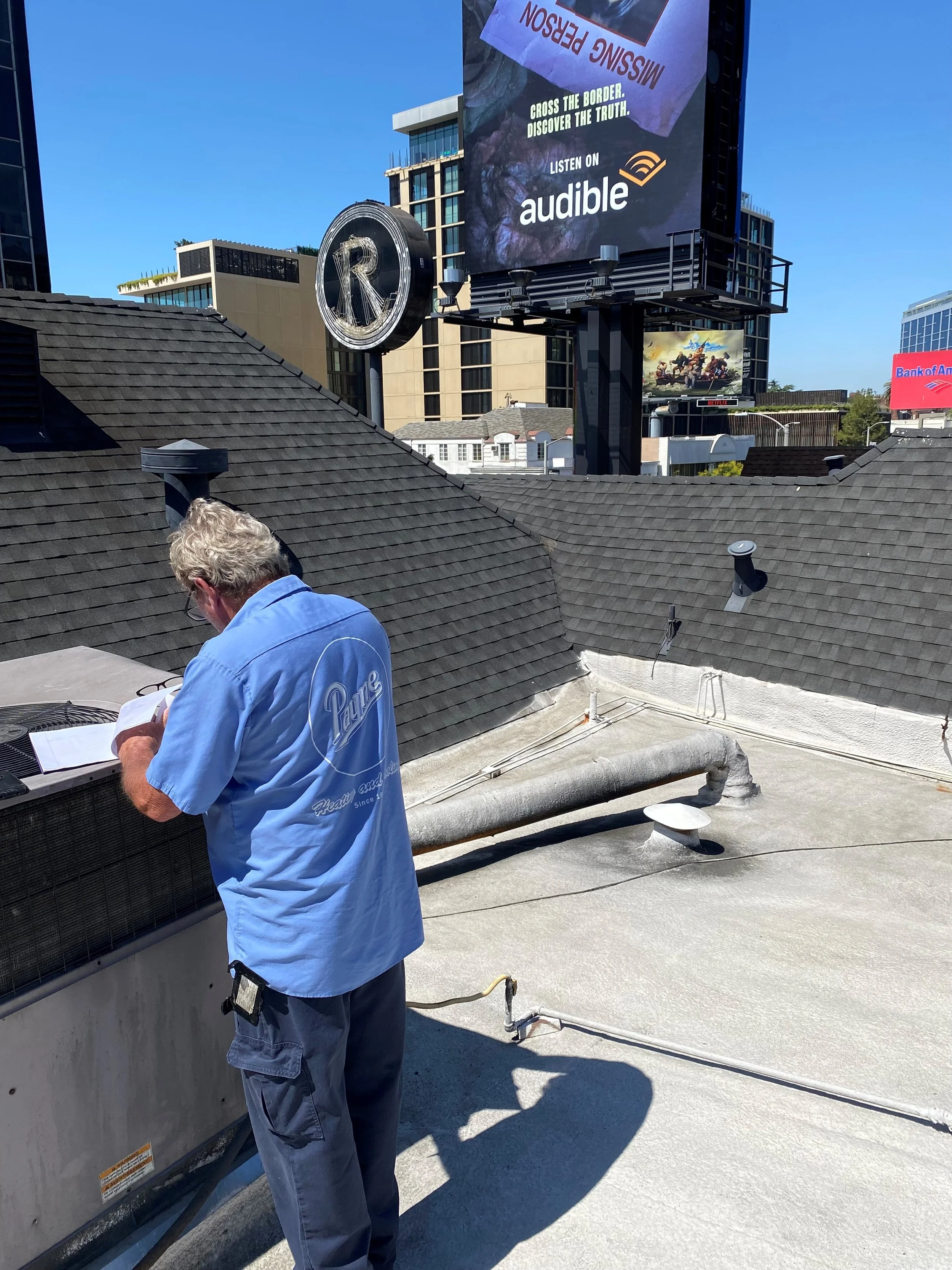 A man with gray hair, wearing a blue shirt and gray pants, is working on a rooftop near an air conditioning unit, with a city and billboard in the background.