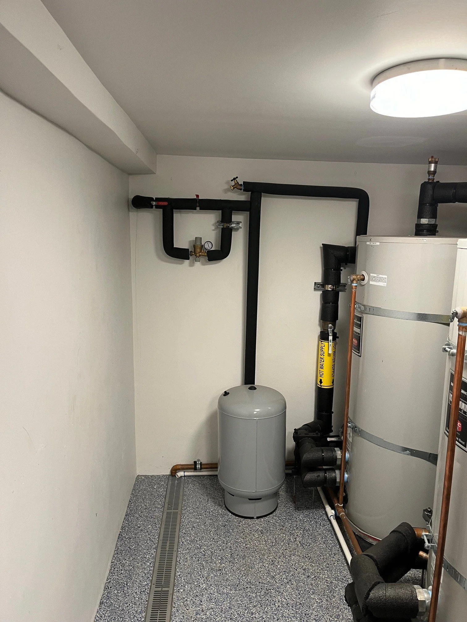Room with water heater, black pipes, copper pipes, pressure gauge, and floor drain.