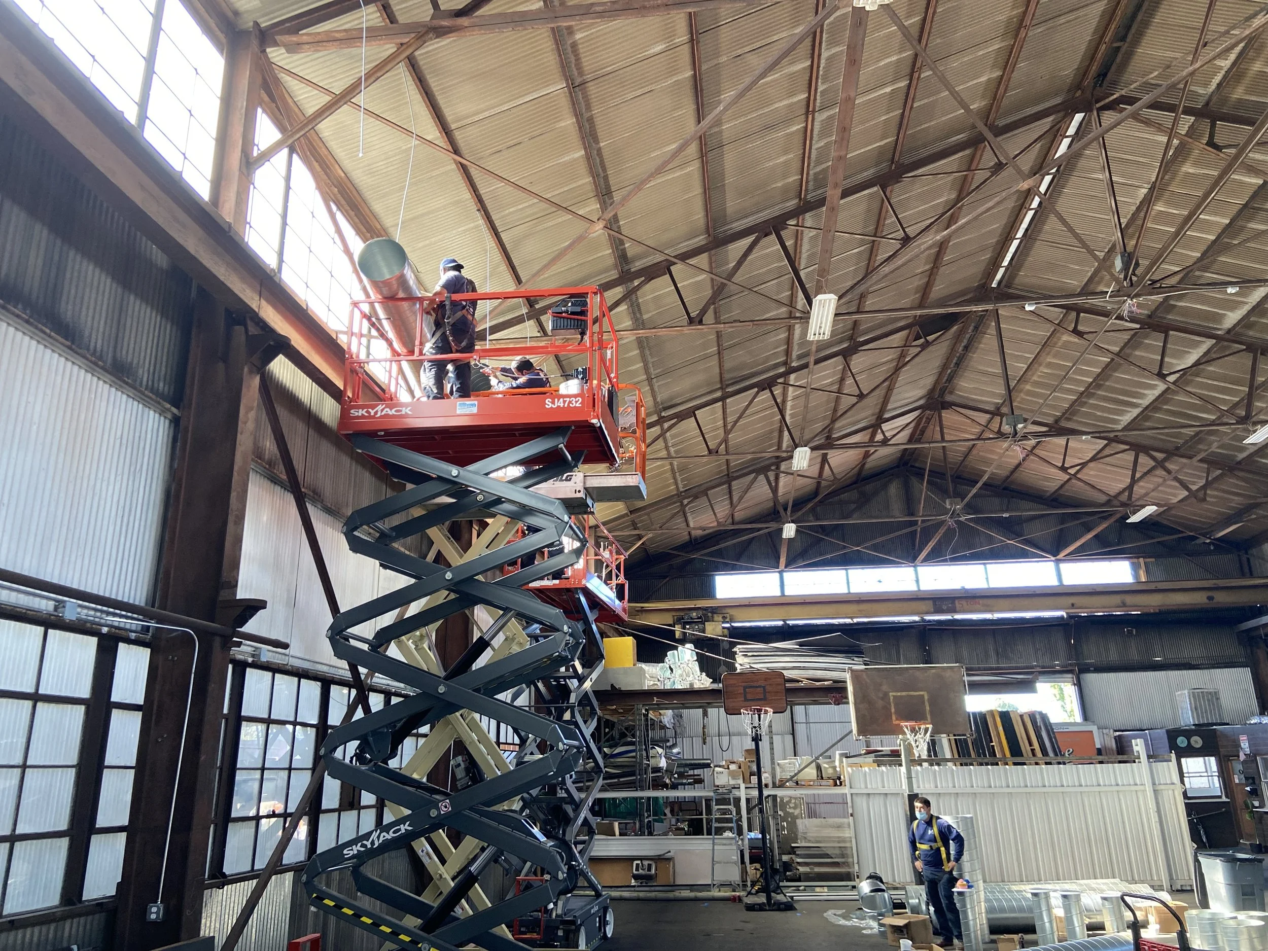 Two workers on a cherry picker lift inside a large warehouse, with additional worker on the ground wearing a mask.