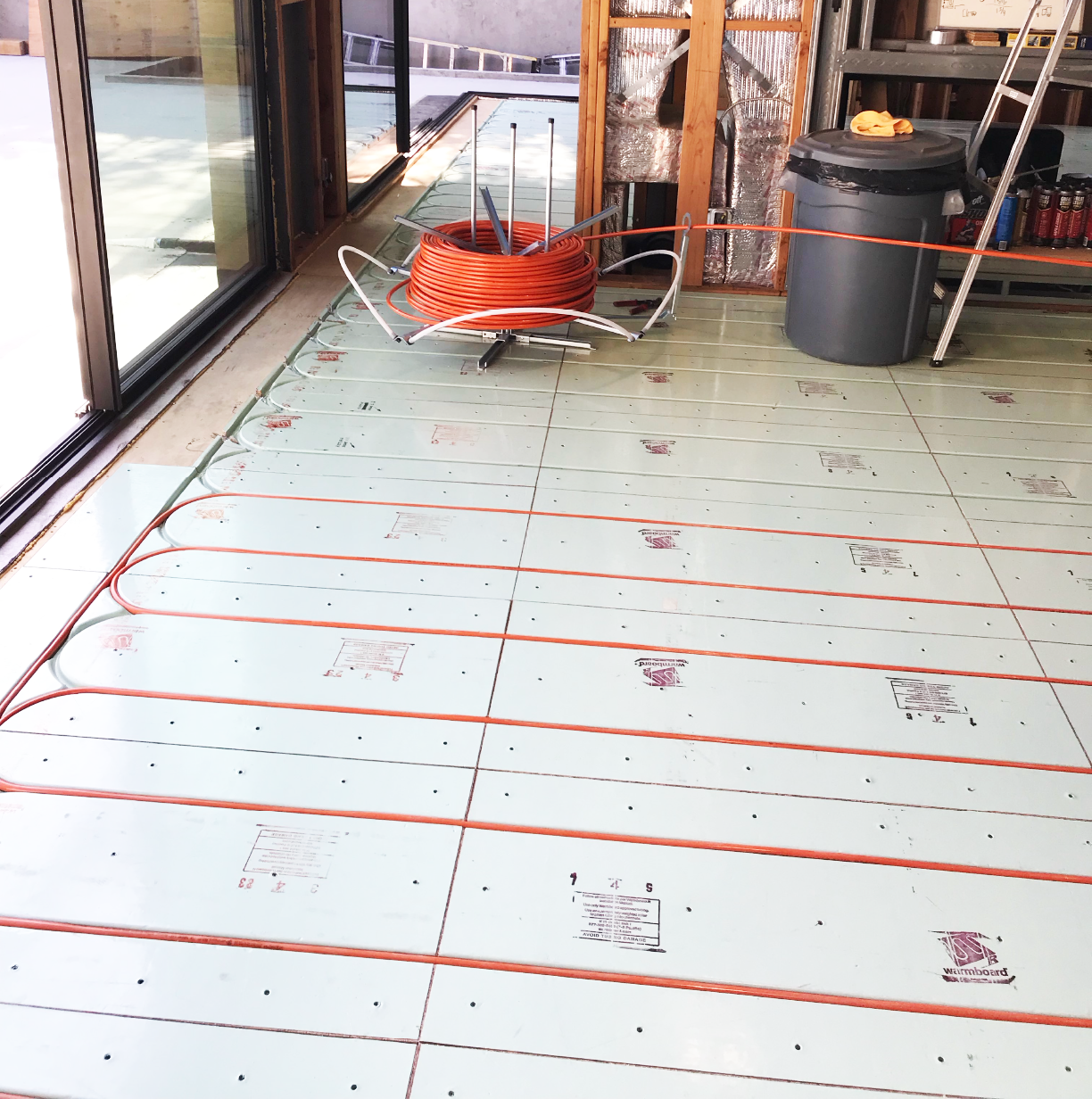 Underfloor radiant heating installation in progress with insulation panels and orange PEX tubing, located inside a partially constructed room with construction tools and materials nearby.