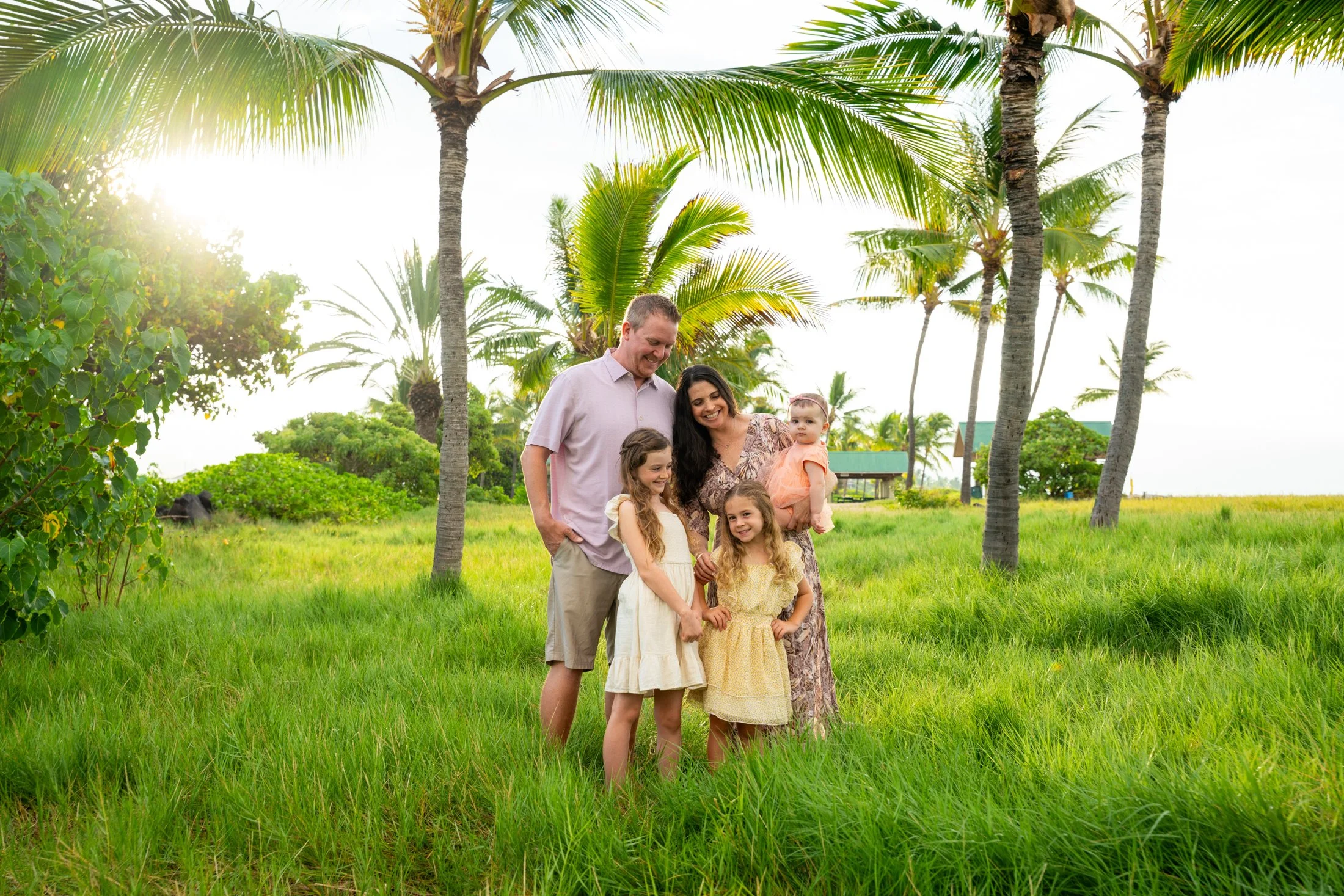 A happy family of five standing on lush green grass in a tropical setting with palm trees, a small house, and bright sunlight in the background.