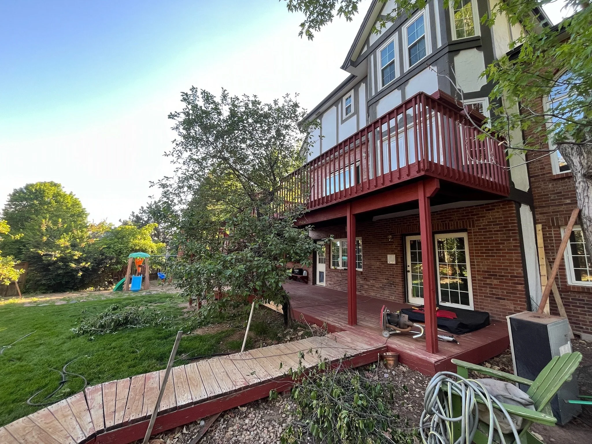 Backyard with a brick and light-colored house, red deck, trees, and a playground in the distance.