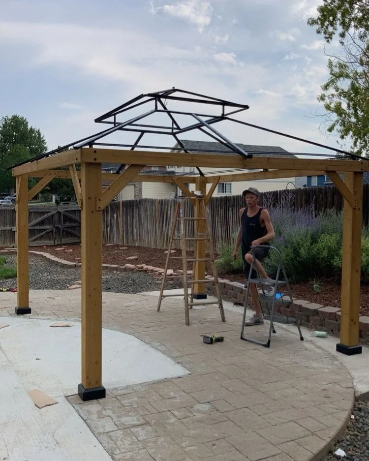 Person working on building a metal frame gazebo in a backyard with wooden structure and stone pathway.