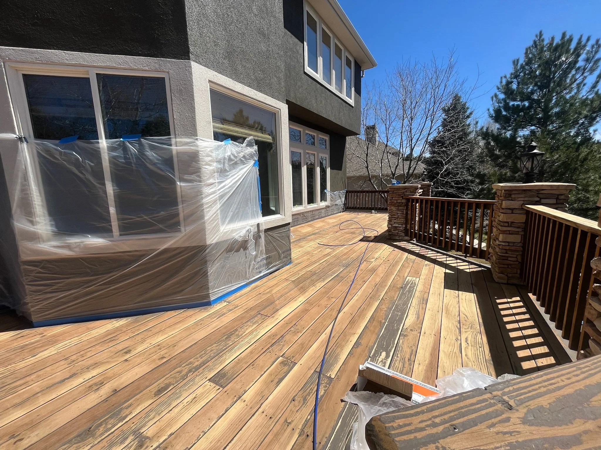 Newly sanded wooden deck attached to house, with some areas still being finished, in a suburban neighborhood with trees and clear blue sky.