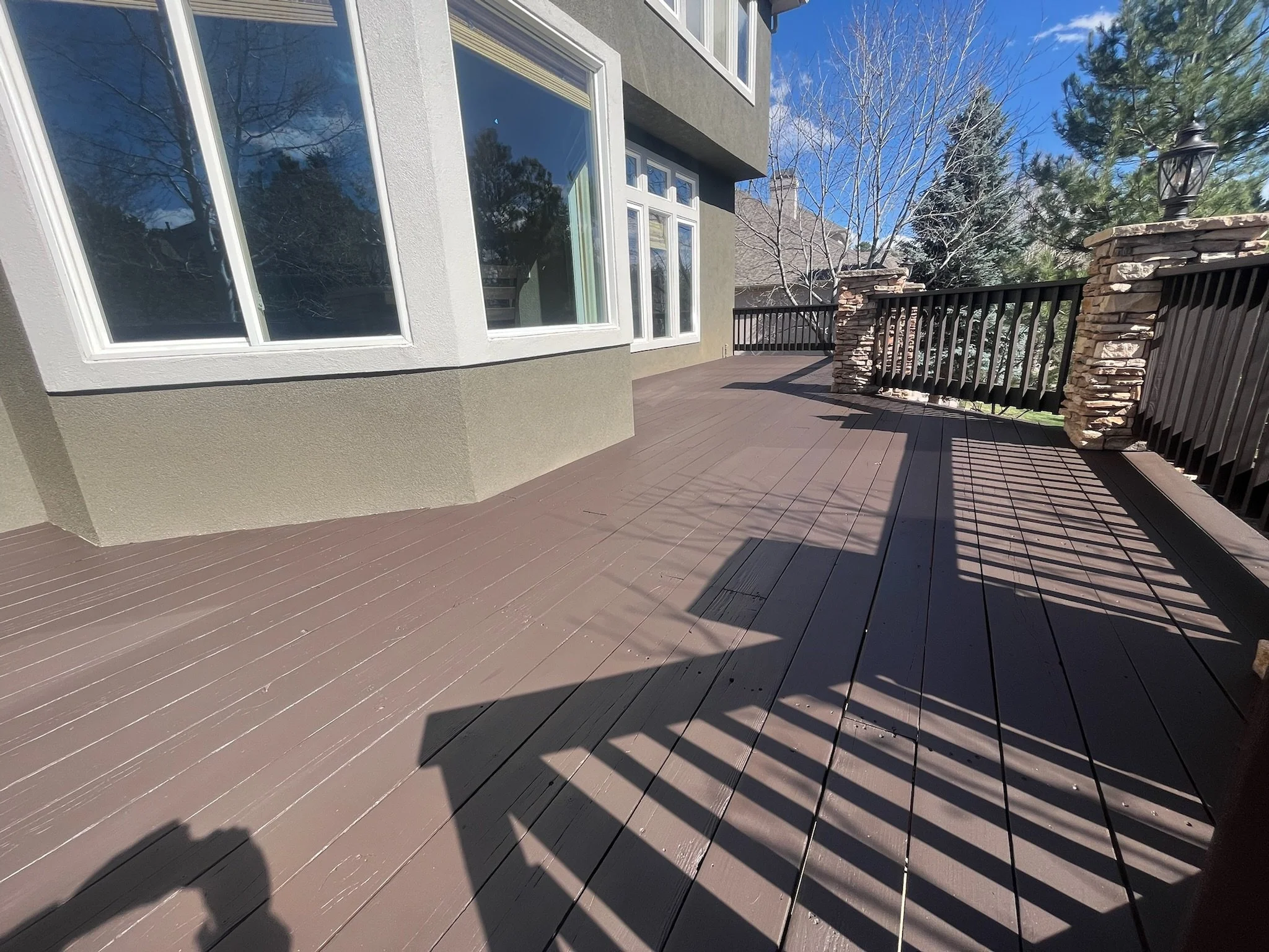 A spacious outdoor wooden deck attached to a house with beige exterior walls and large white-framed windows. The deck has a black metal railing with stone pillars, and there are trees in the background under a blue sky.