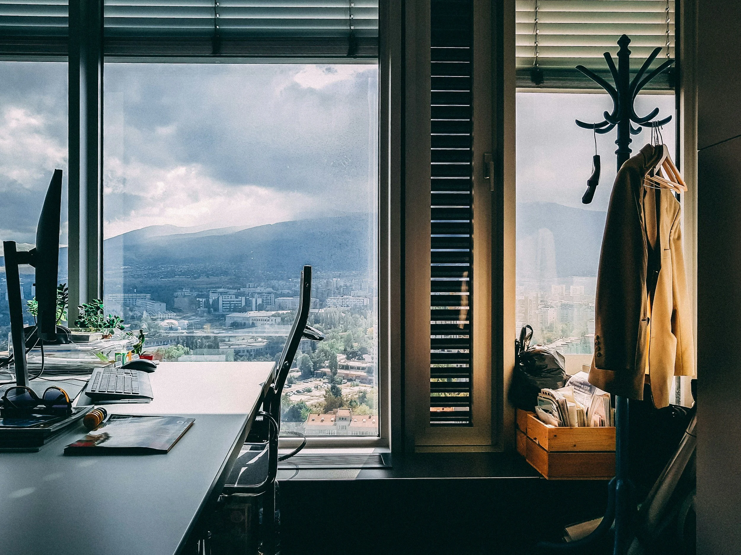Office room with large window overlooking city and mountains, desk with computer and office supplies, coat rack with coat and hangers, wooden crate with magazines and bags.