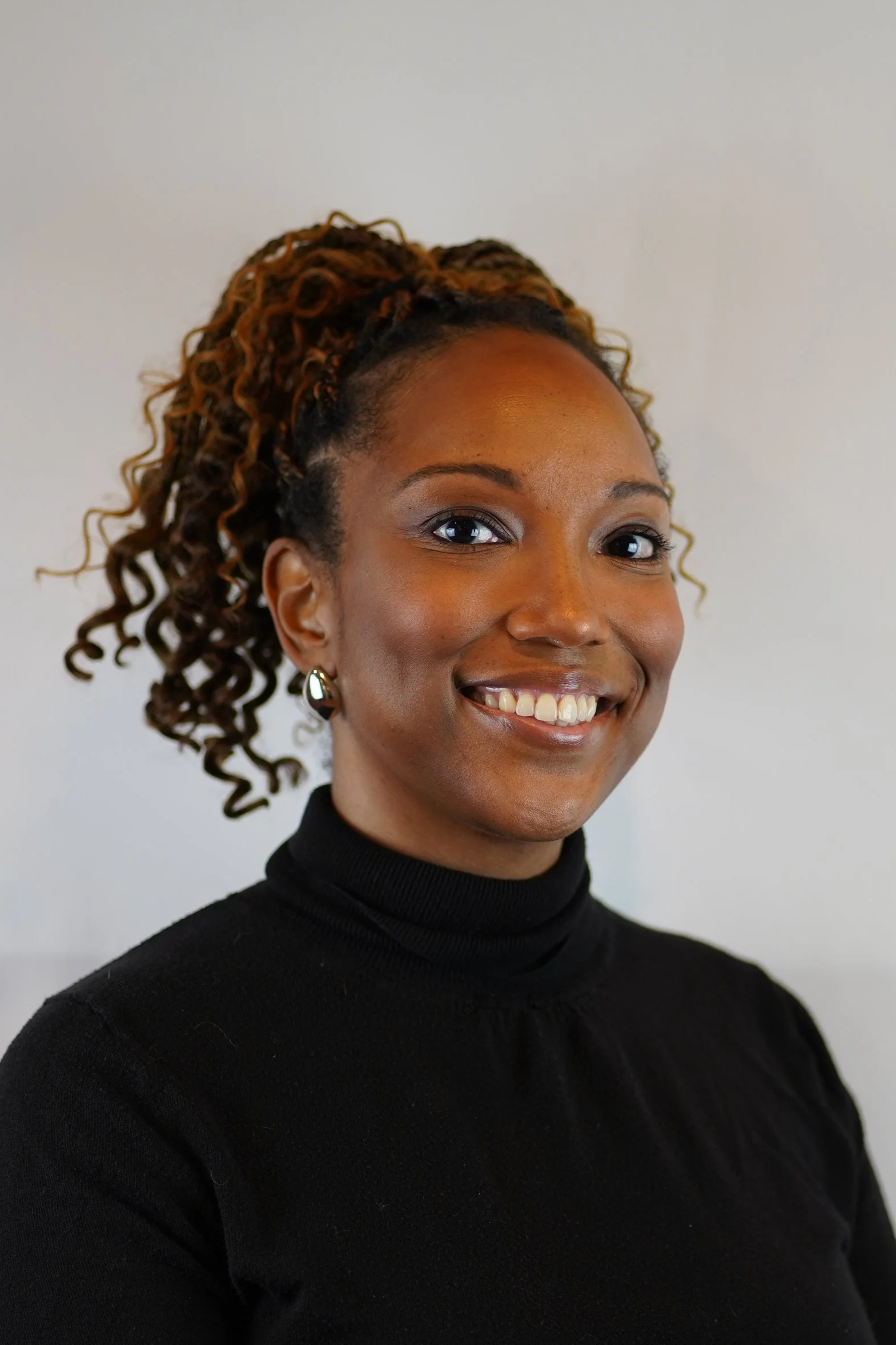 Portrait of a smiling woman with curly hair in a high ponytail, wearing a black turtleneck and earrings, against a plain light background.