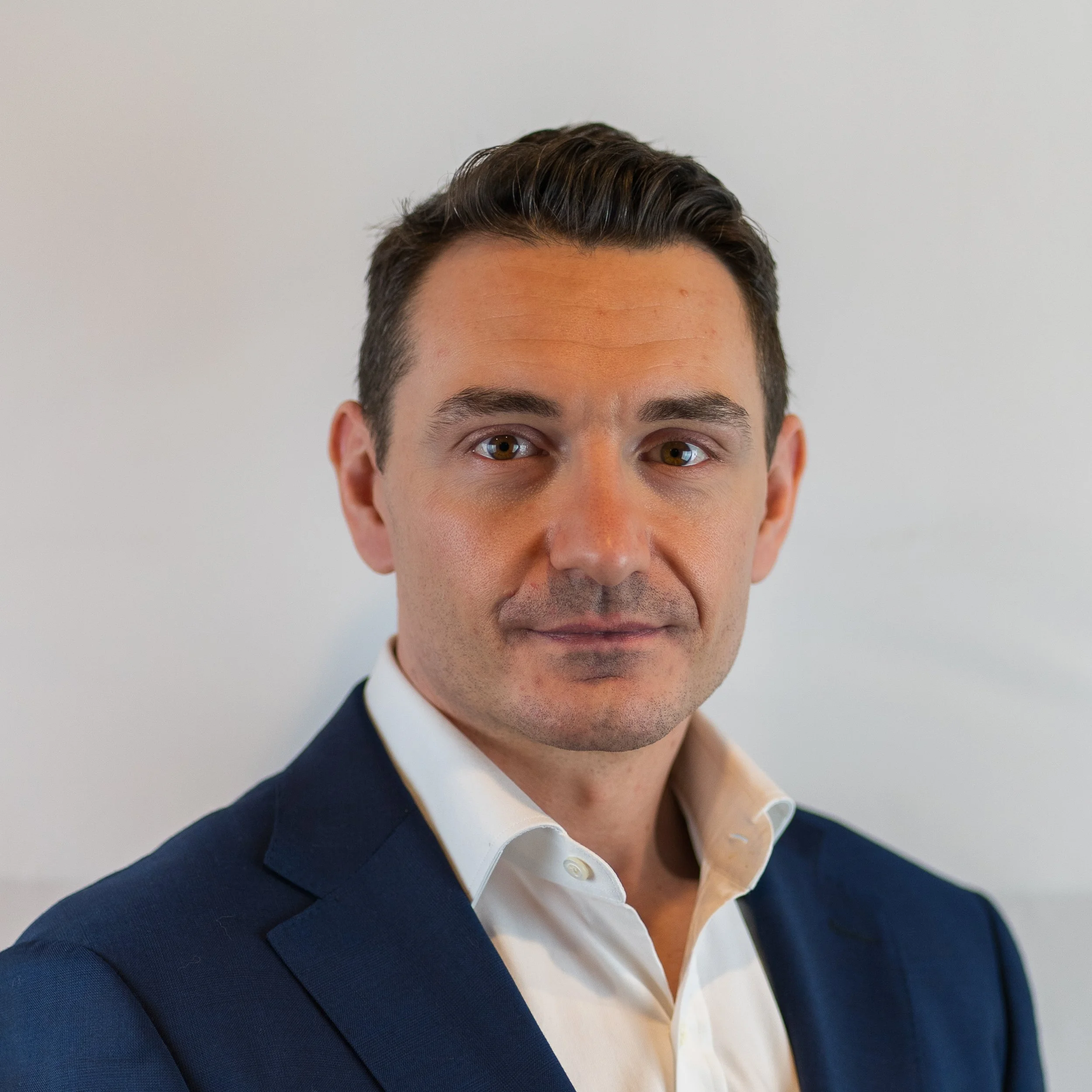 A professional headshot of a man in a dark blue suit and white shirt, with short dark hair and light skin, standing against a plain background.