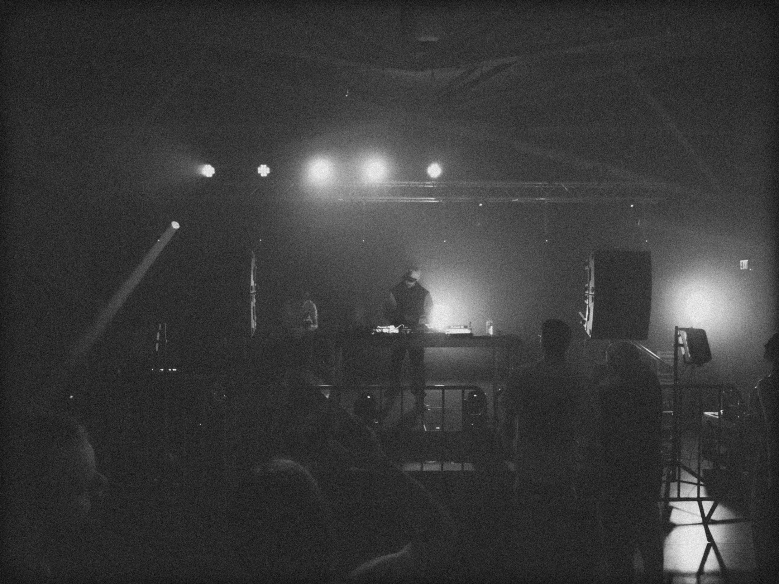 Flippin’ with intense expression, mid-transition during a live tech house DJ set, surrounded by cables and gear