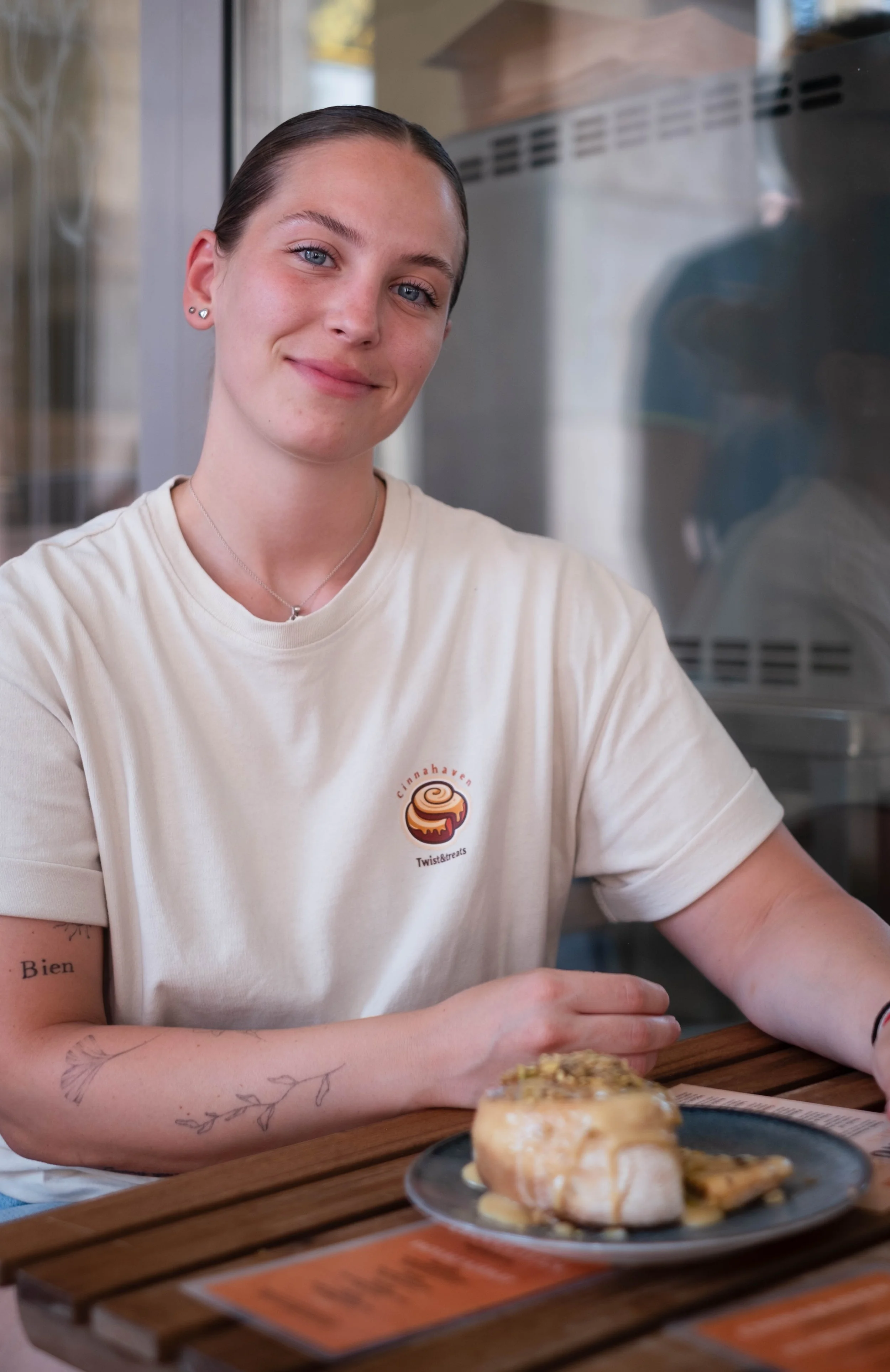 A young woman with blue eyes and dark hair tied back, smiling at the camera, wearing a beige T-shirt with a cinnamon roll logo. She is sitting at a wooden table with a plate of dessert that includes a cinnamon roll topped with icing and nuts. The background shows a glass window and a refrigerator or oven.