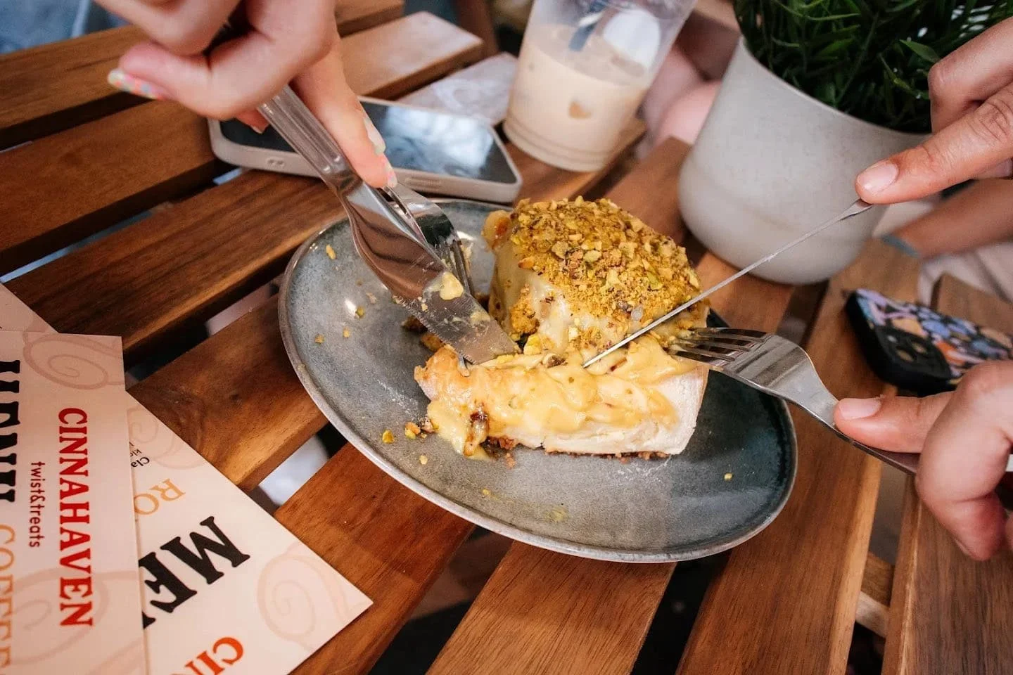 A slice of cheesecake topped with crushed pistachios on a gray plate, with three people cutting and sharing it at a wooden table. A smartphone, a drink, and a plant are visible on the table.