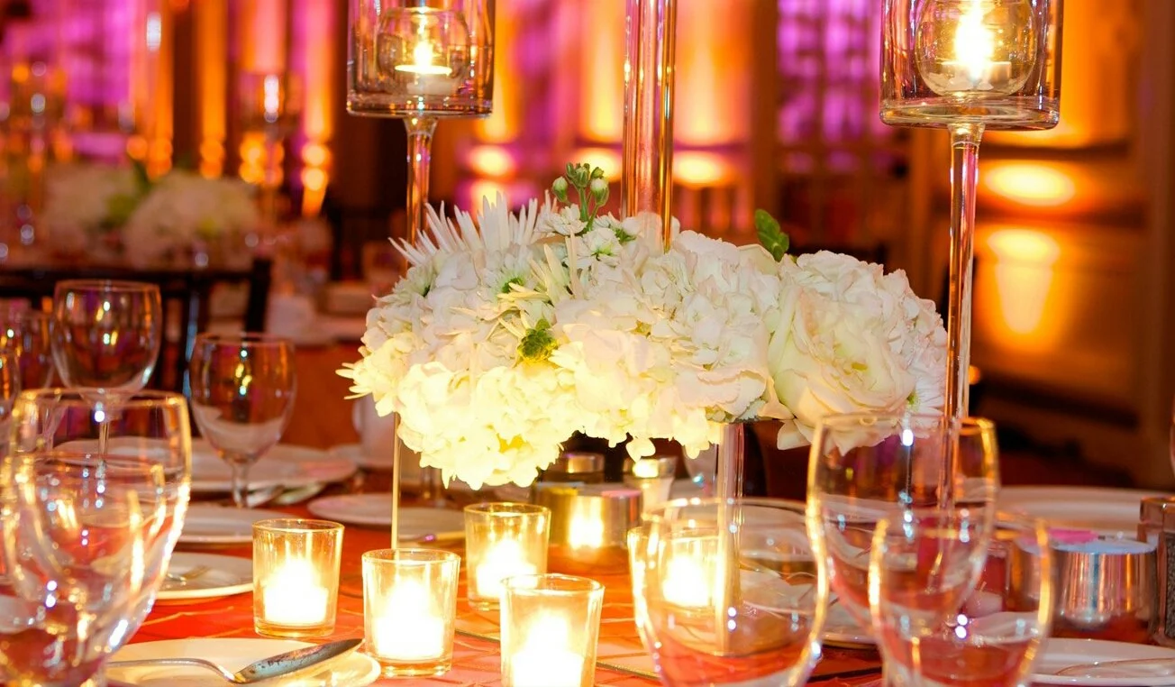 Elegant table setting with a floral centerpiece of white flowers, surrounded by candles, wine glasses, and tall glass candle holders at a formal event or wedding reception.
