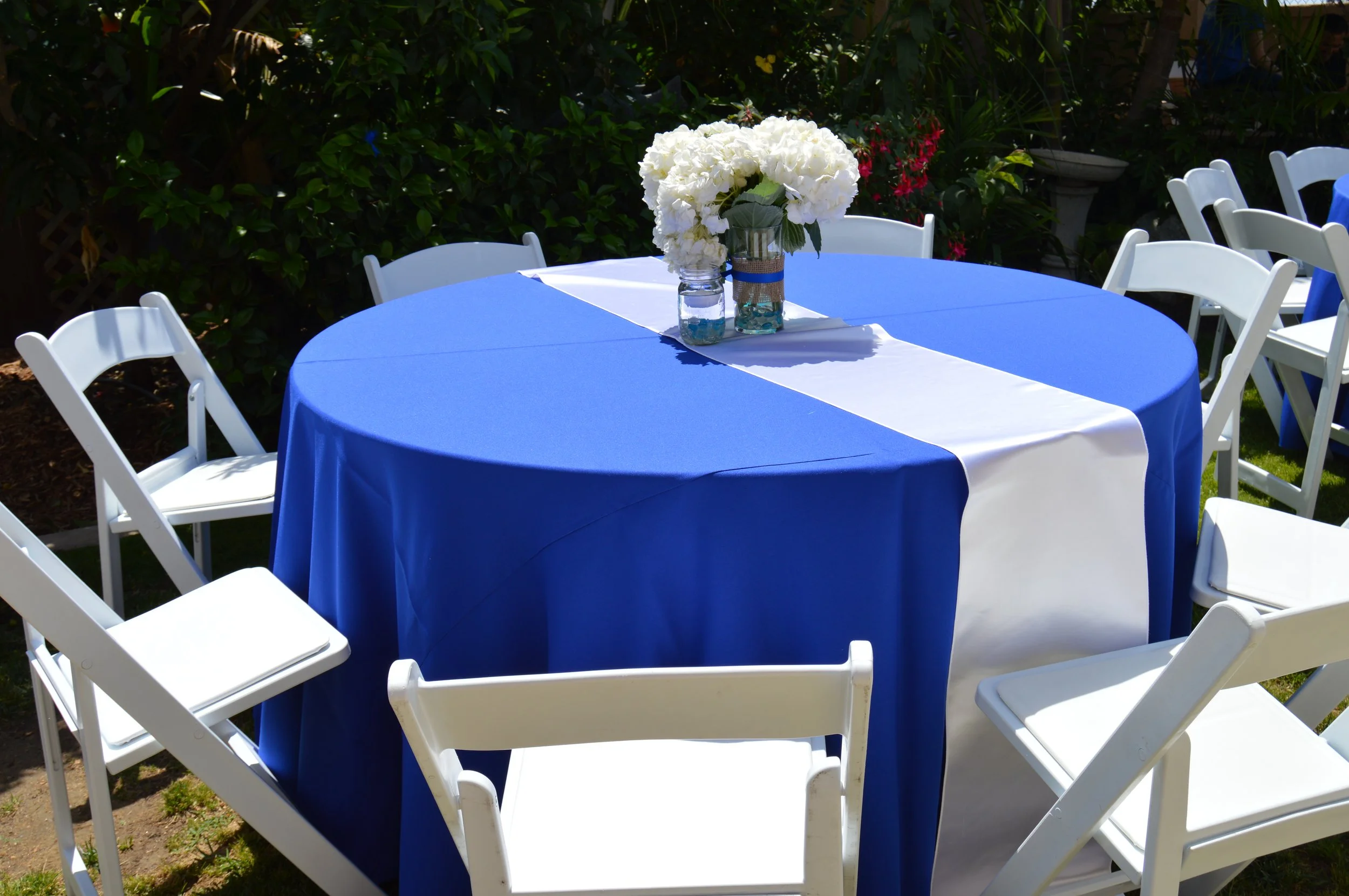 Round table with a blue tablecloth and a white table runner, decorated with a bouquet of white hydrangeas in glass vases, set outdoors surrounded by white chairs and lush greenery.