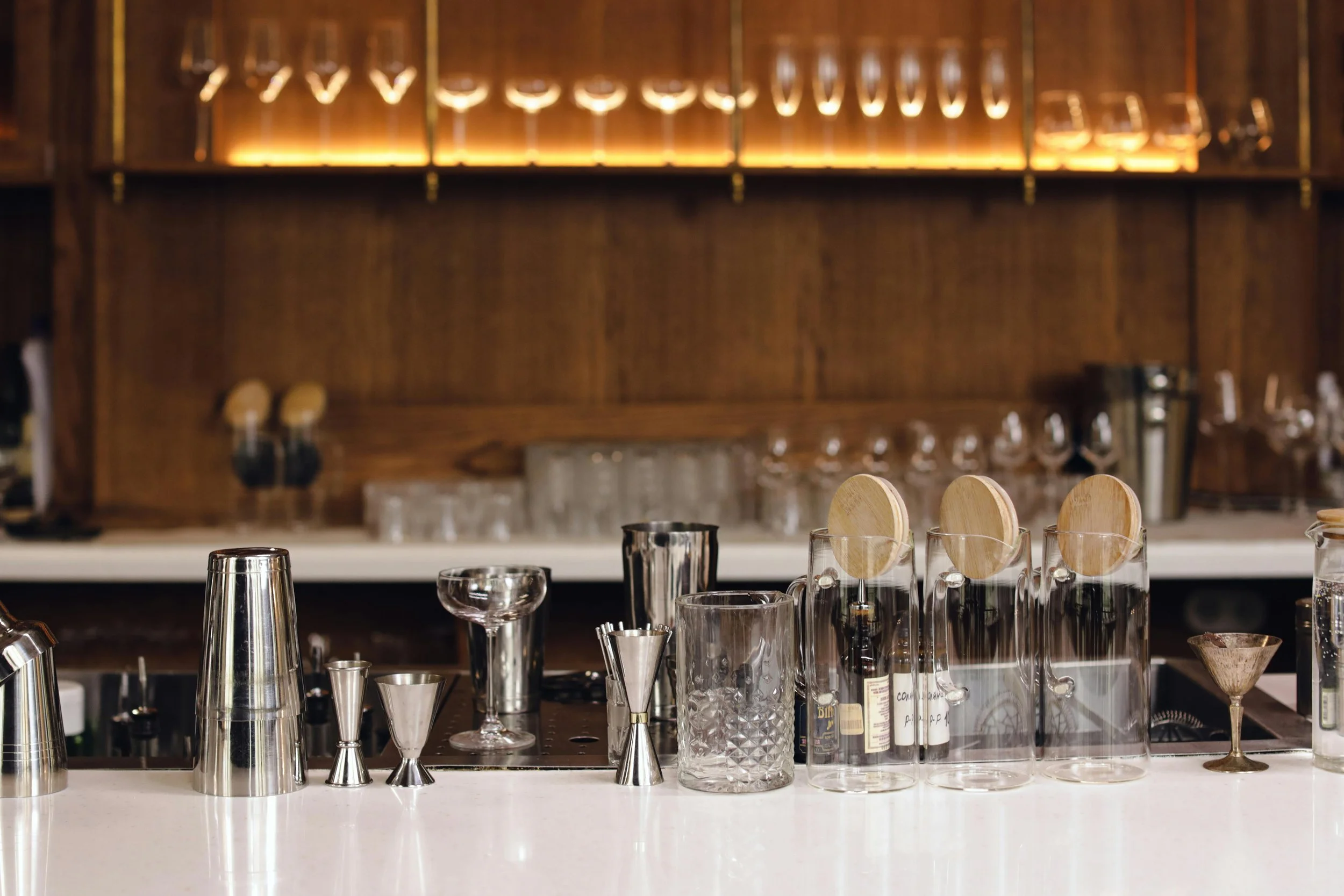 Bar counter with various cocktail-making tools including shakers, jiggers, and glasses, with a wooden shelf stocked with glasses and barware in the background.
