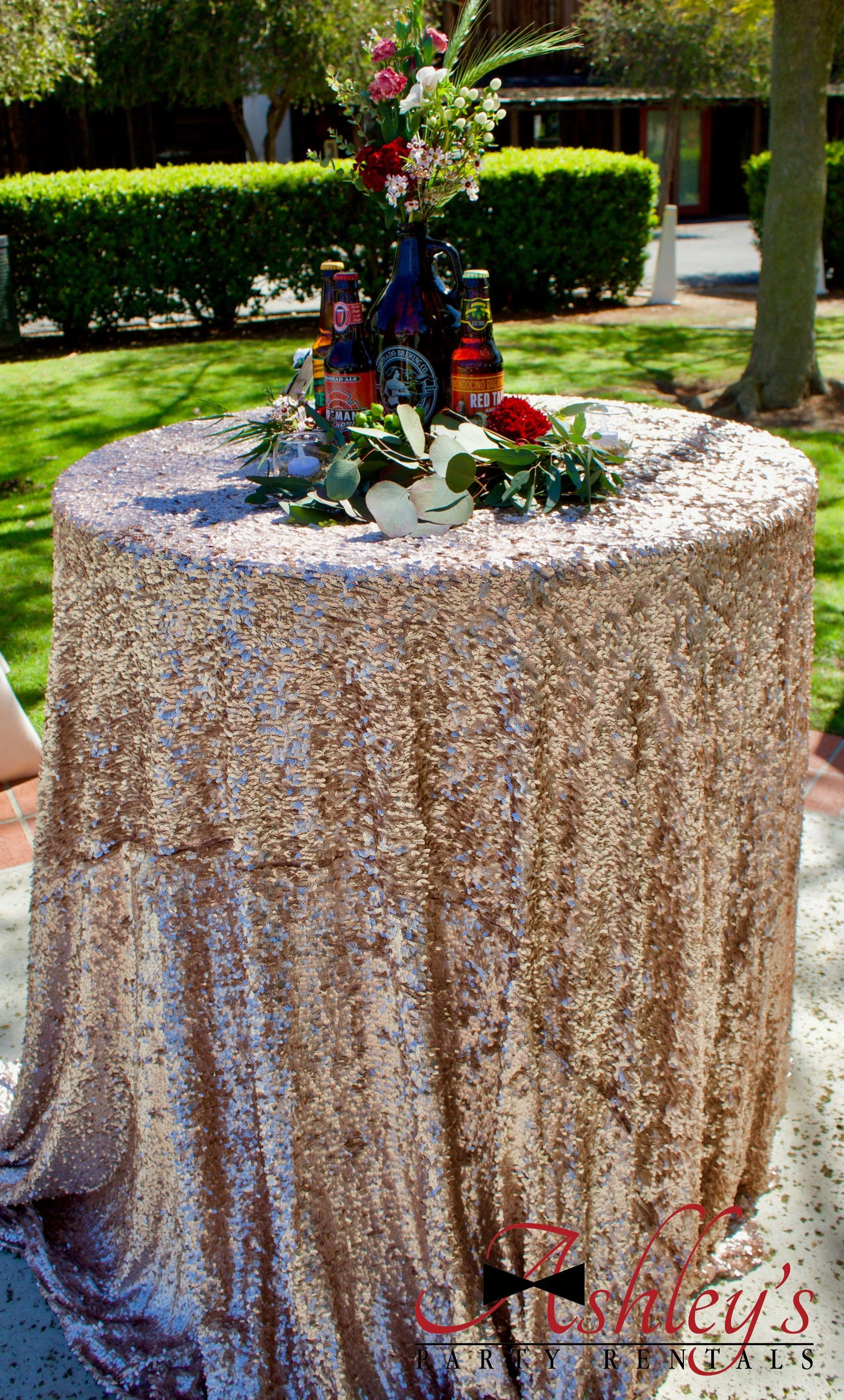 Round table covered with a sparkly, textured gold tablecloth, topped with a floral arrangement in a dark blue pitcher, surrounded by small flowers, leaves, and two beer bottles. In the background, there are trees and a hedge outdoors.