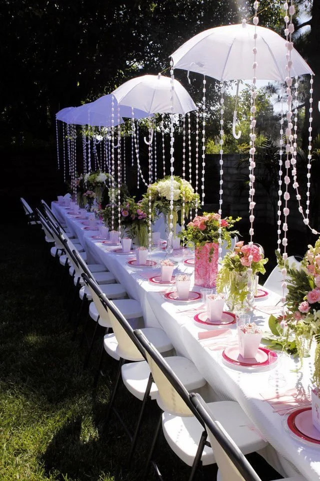 Outdoor event table decorated with pink and white flowers, umbrellas, and hanging beads, set for a celebration or gathering.
