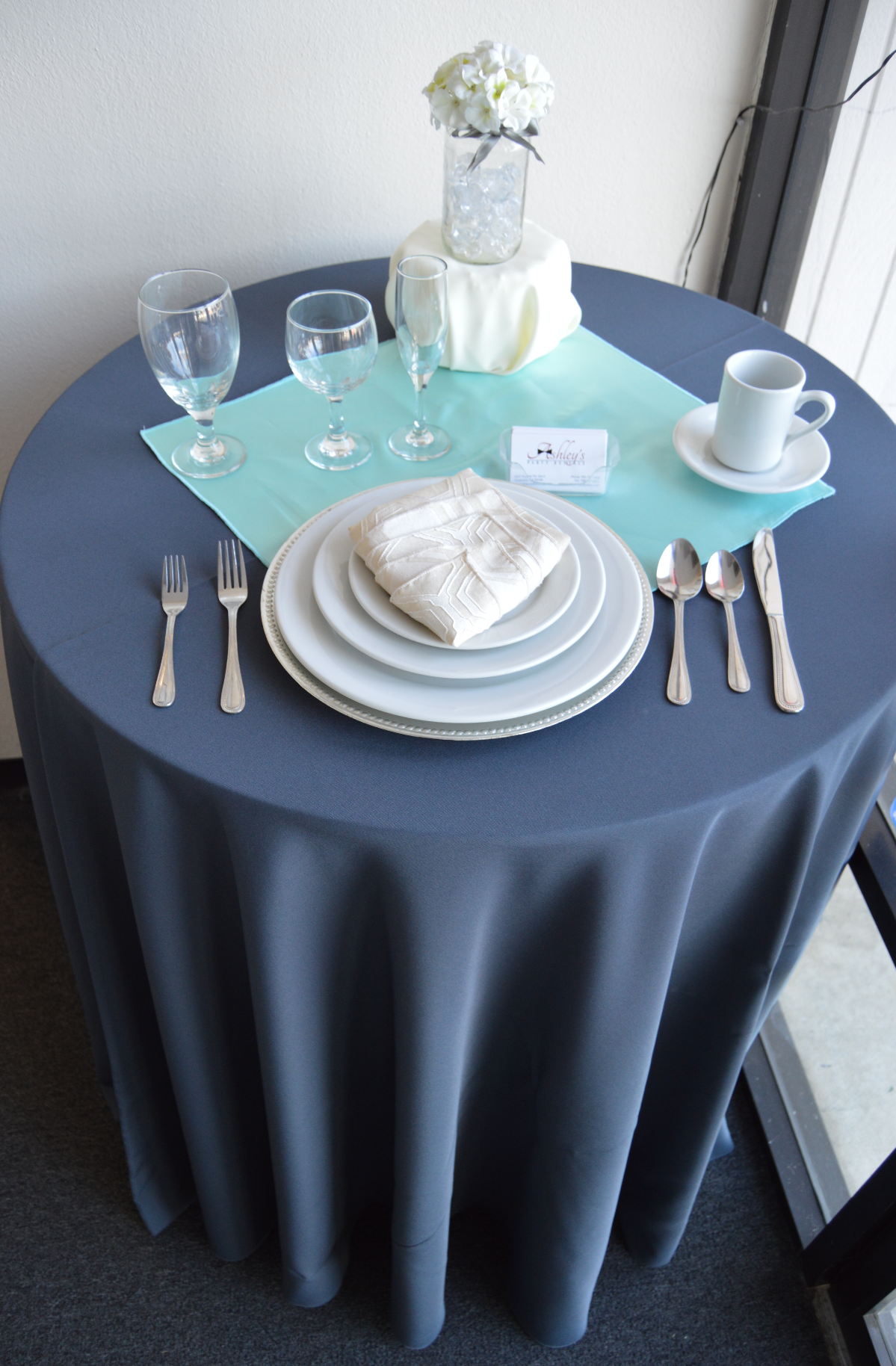 Elegant round table setup with a dark blue tablecloth, white plates, napkin, cutlery, wine glasses, a white coffee mug on a saucer, a light blue placemat, a small card, and a glass vase with white flowers.