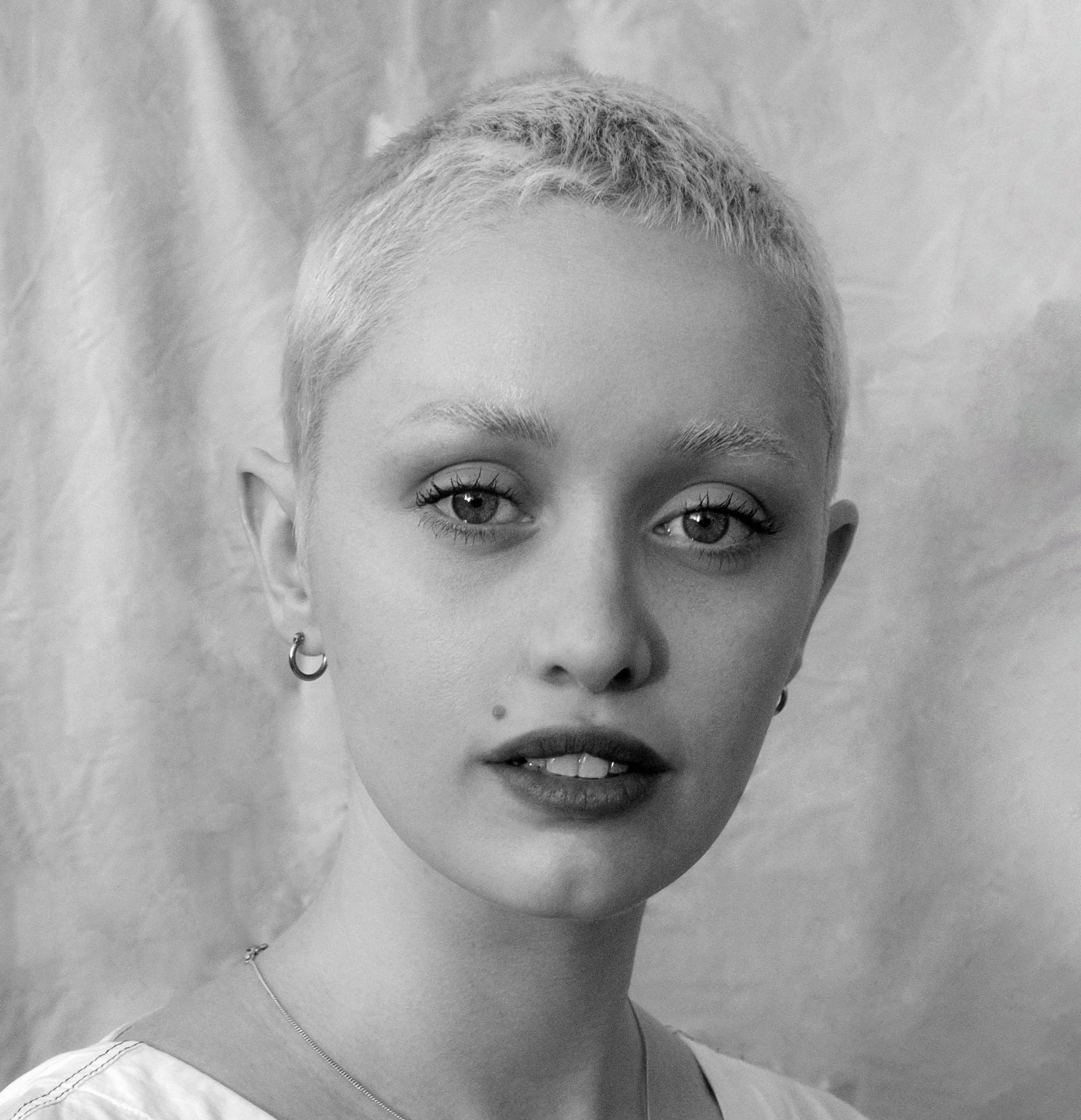 Close-up black and white photo of a young woman with short, light-colored hair, wearing earrings, a necklace, and a jacket, with a blurred background.