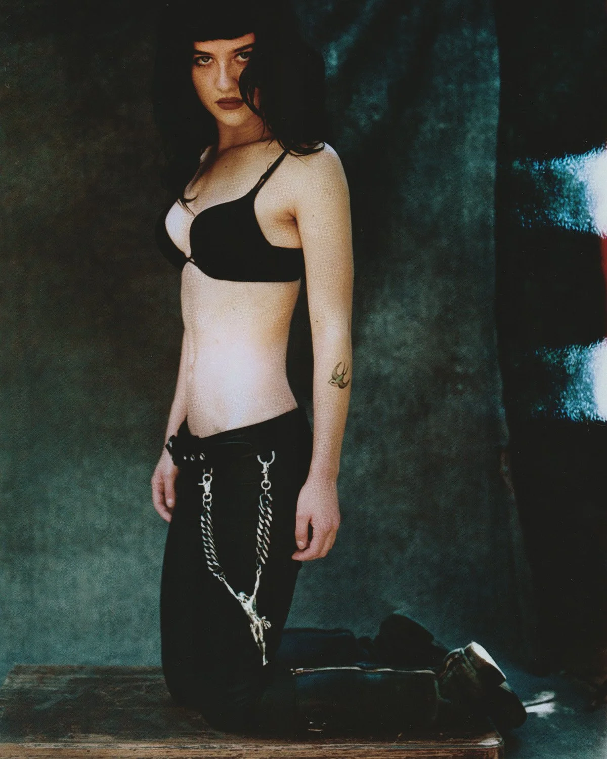 A woman with black hair and dark lipstick kneeling on a wooden surface against a dark background, wearing a black bra and black pants with chains hanging from the waistband, with a tattoo on her left forearm.