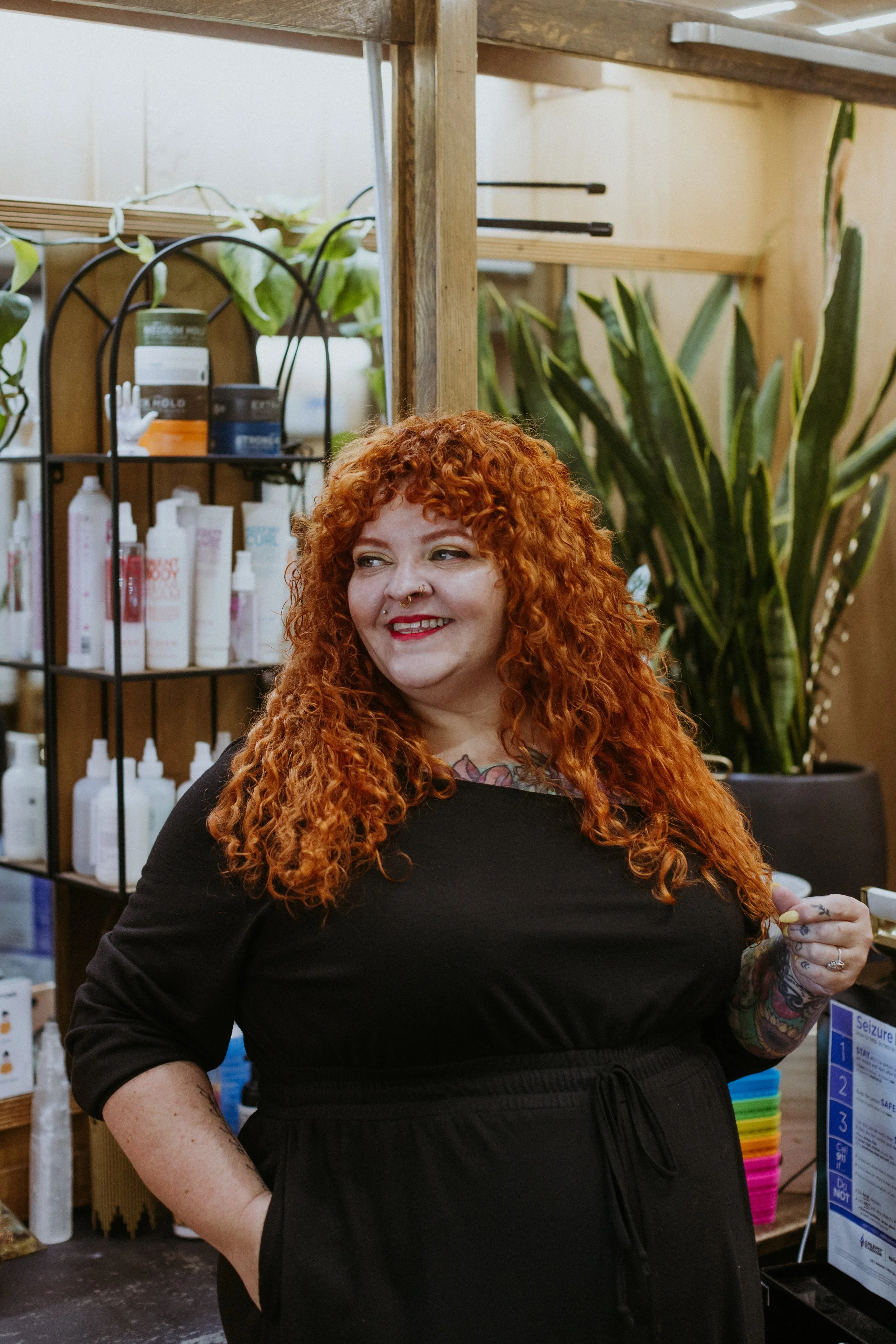 A smiling woman with curly red hair and tattoos on her arm wearing a black top and pants, standing in a store with shelves of beauty products and green plants in the background.