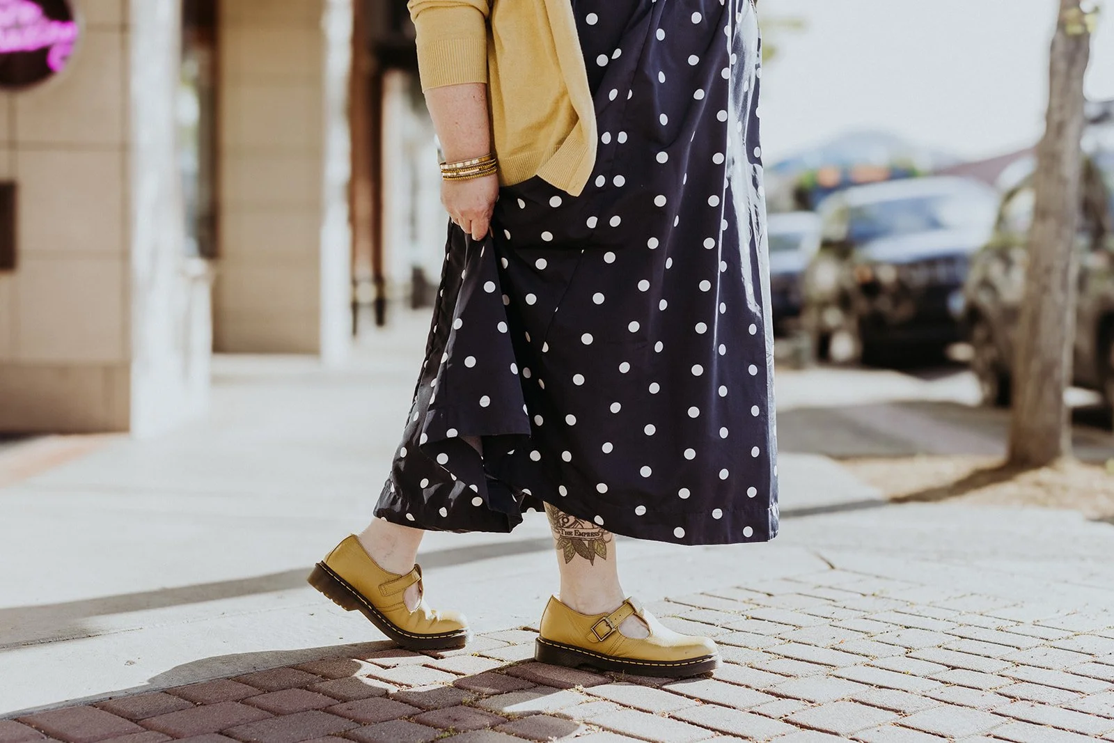 Person walking on brick sidewalk, wearing a navy blue polka dot dress, yellow shoes, a yellow cardigan, and gold bracelets, with tattoo on ankle visible.
