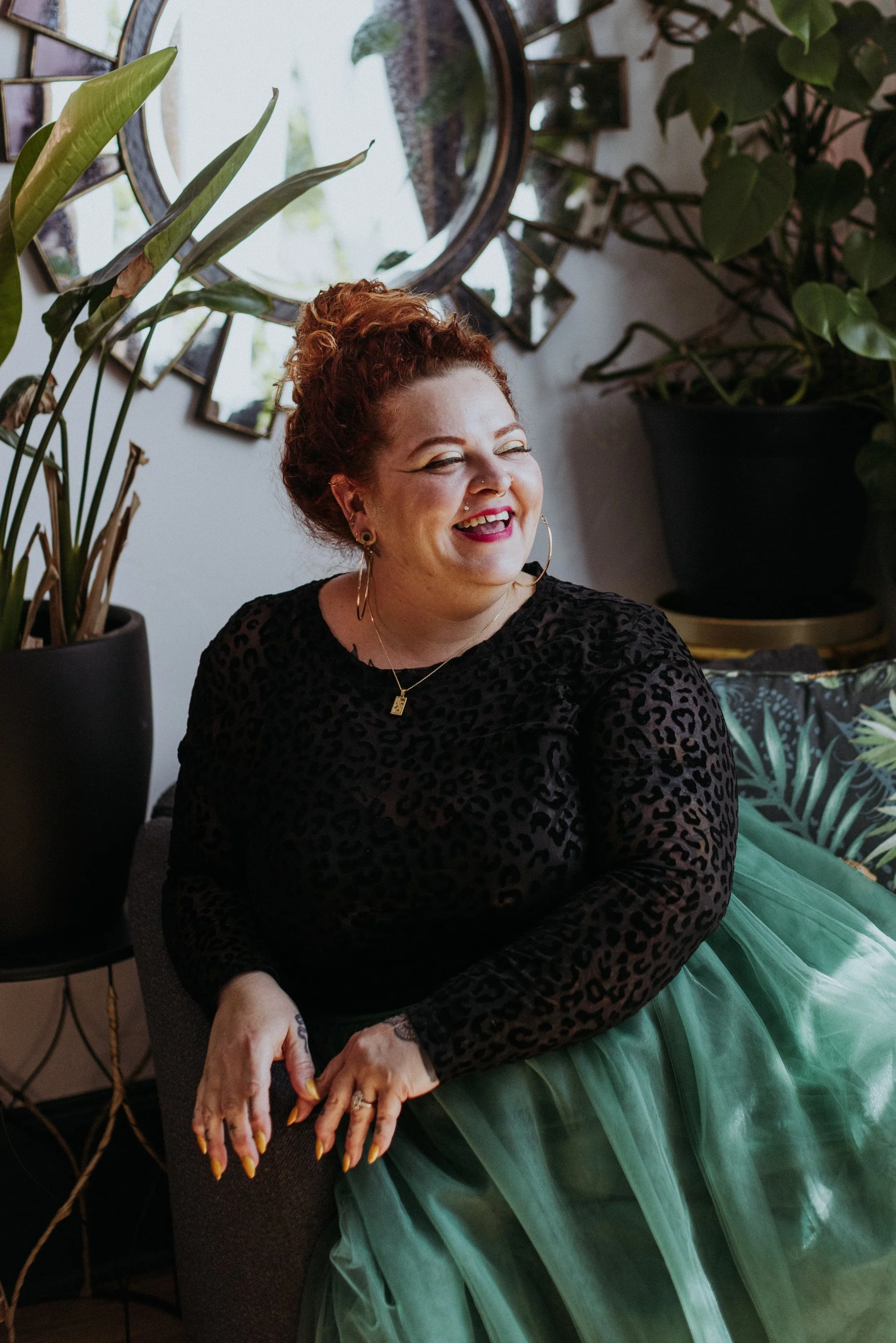 A woman with curly red hair styled in a bun, wearing a black leopard print top, gold hoop earrings, a gold necklace, and bright lipstick, sitting indoors near a large leafy plant and a mirror, smiling and enjoying herself.