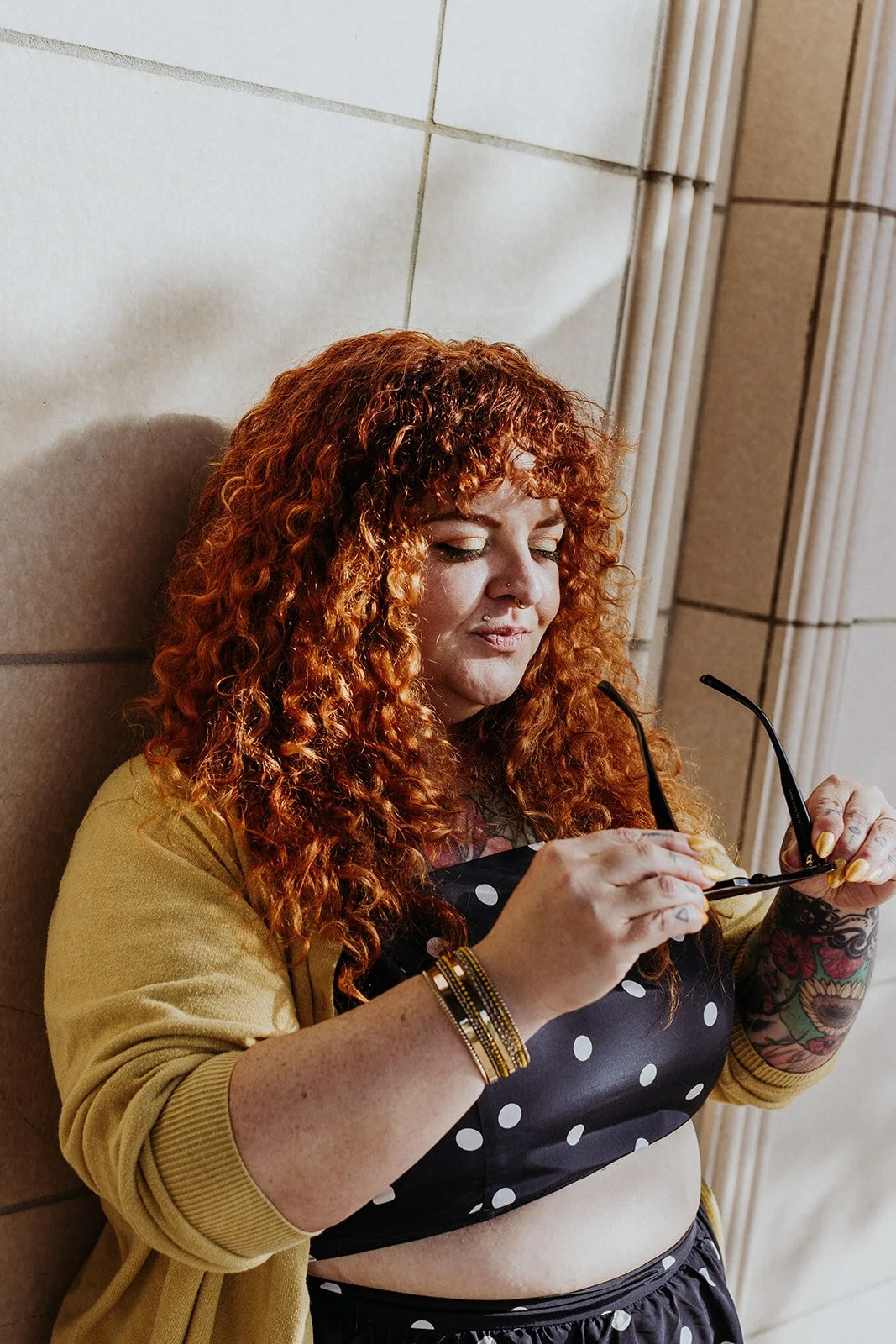 A woman with curly red hair, wearing a yellow jacket and a black polka dot outfit, is sitting against a tiled wall, adjusting her sunglasses with her eyes closed.