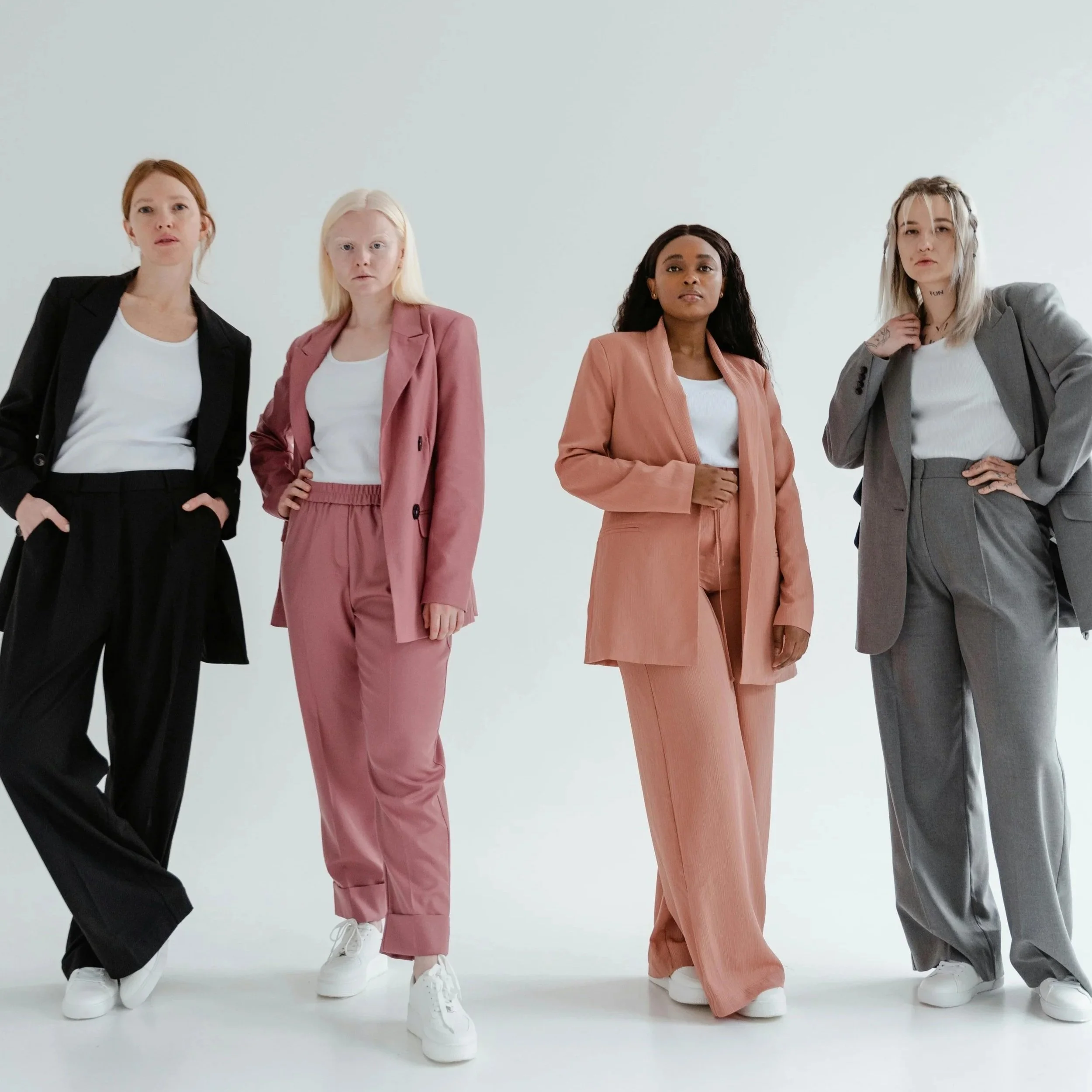 Four women standing together in stylish suits and white sneakers against a plain white background.