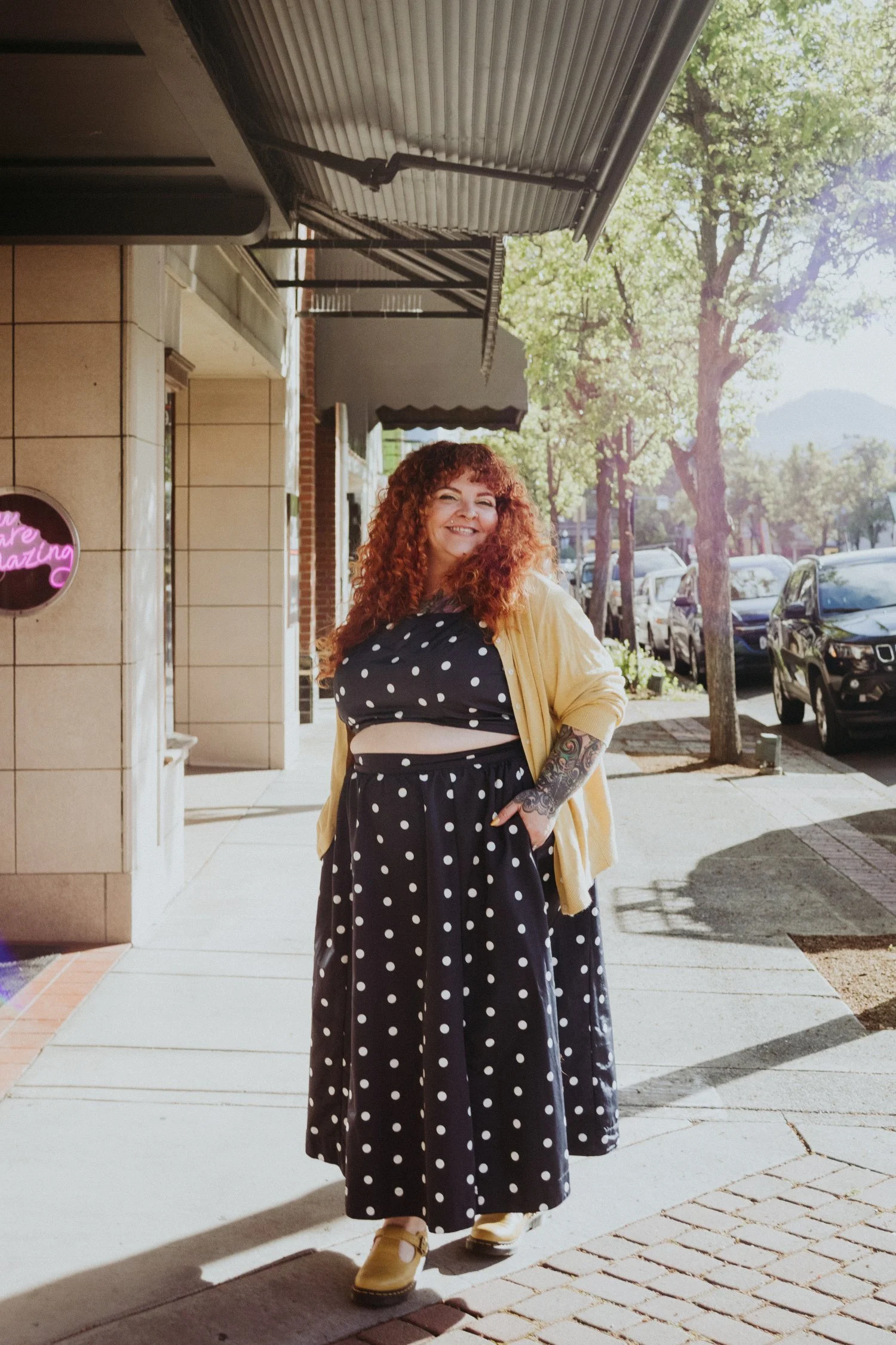 A woman with curly red hair, wearing a polka dot dress, a yellow jacket, and tan shoes, smiling and walking on a sidewalk in front of storefronts and parked cars, with trees and mountains in the background.