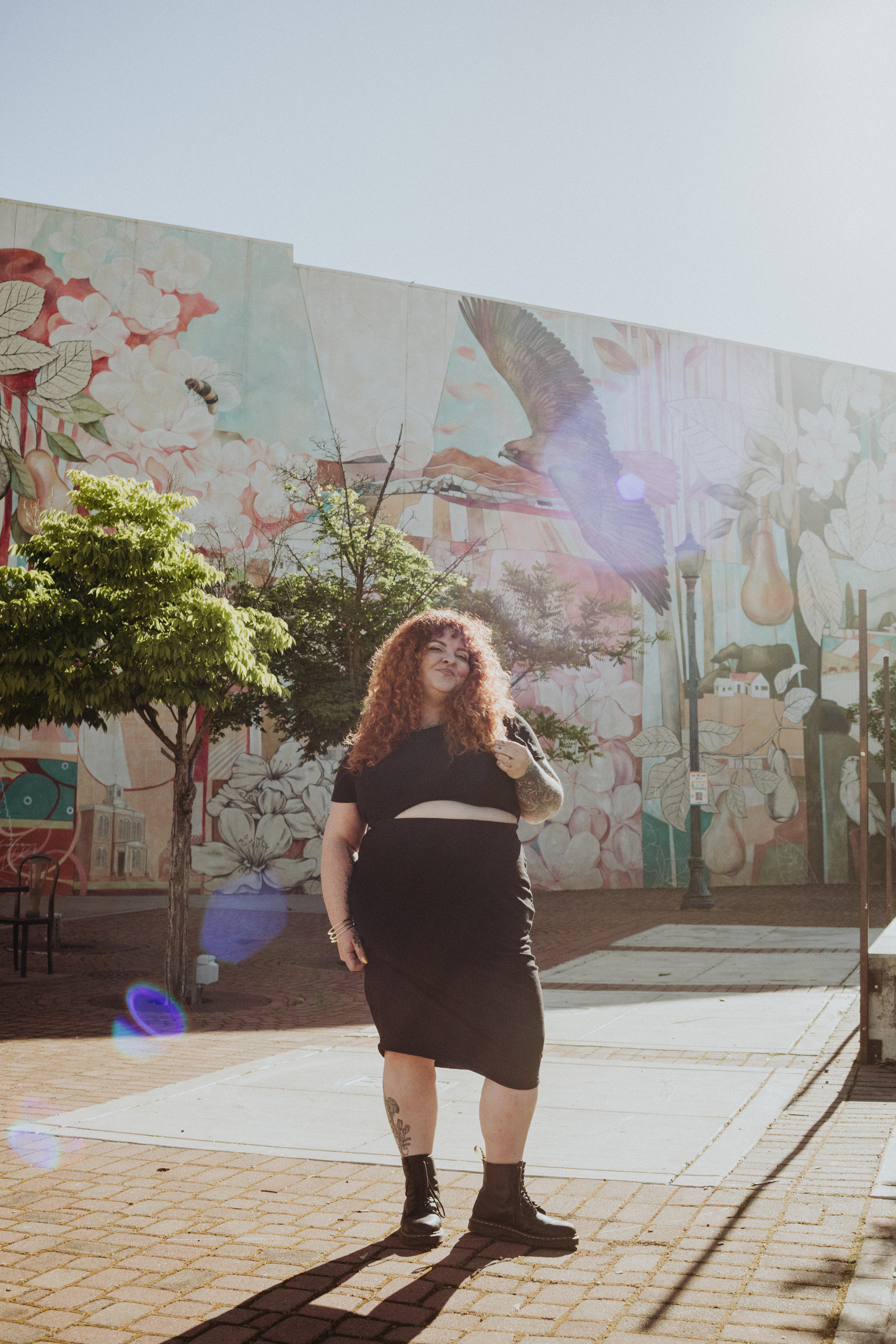 A woman with curly red hair, wearing a black dress, standing on a sunny sidewalk with a colorful mural in the background and a tree nearby.