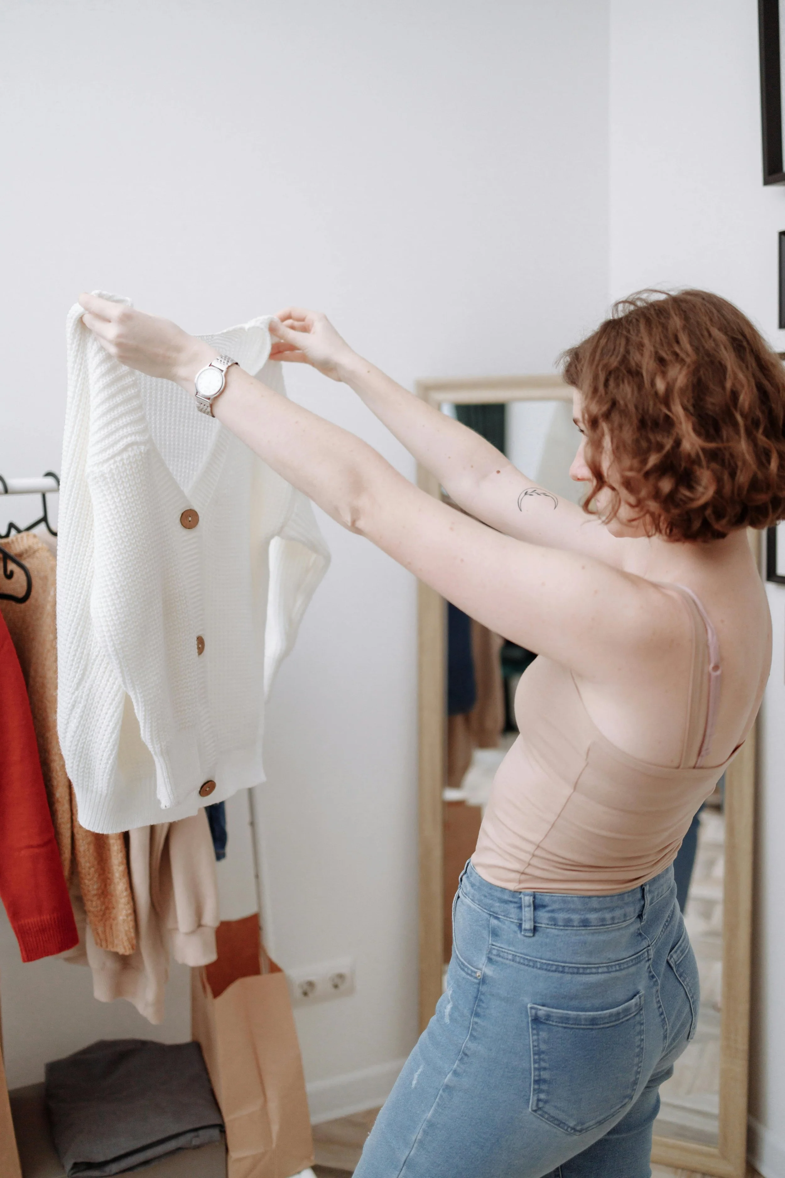 A woman with curly red hair, wearing a beige tank top and blue jeans, is hanging a cream-colored cardigan on a clothing rack.