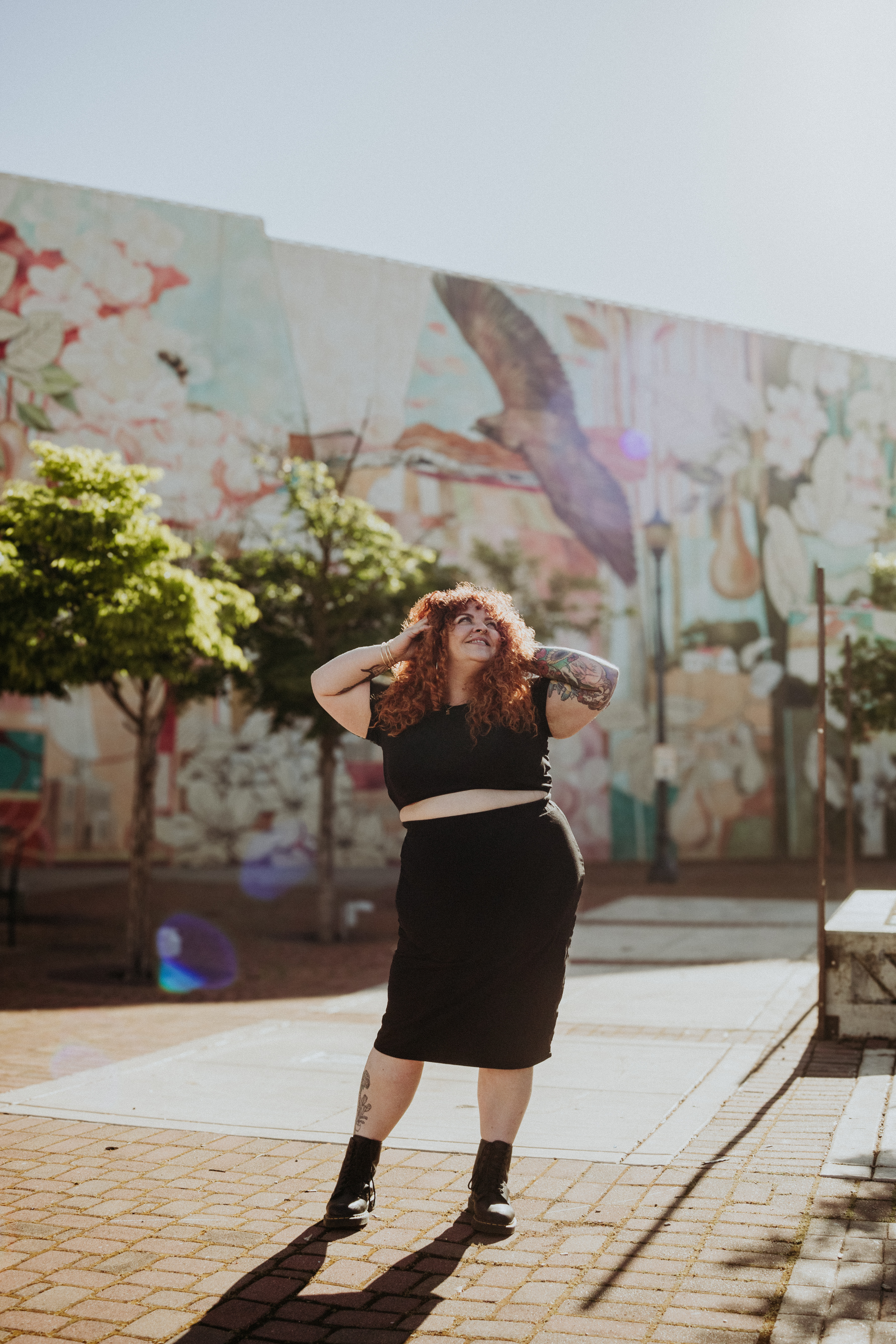 A woman with curly red hair and tattoos on her left arm and right leg, wearing a black crop top, black skirt, and black boots, standing outdoors on a sunny day with a colorful mural, trees, and sidewalk in the background.
