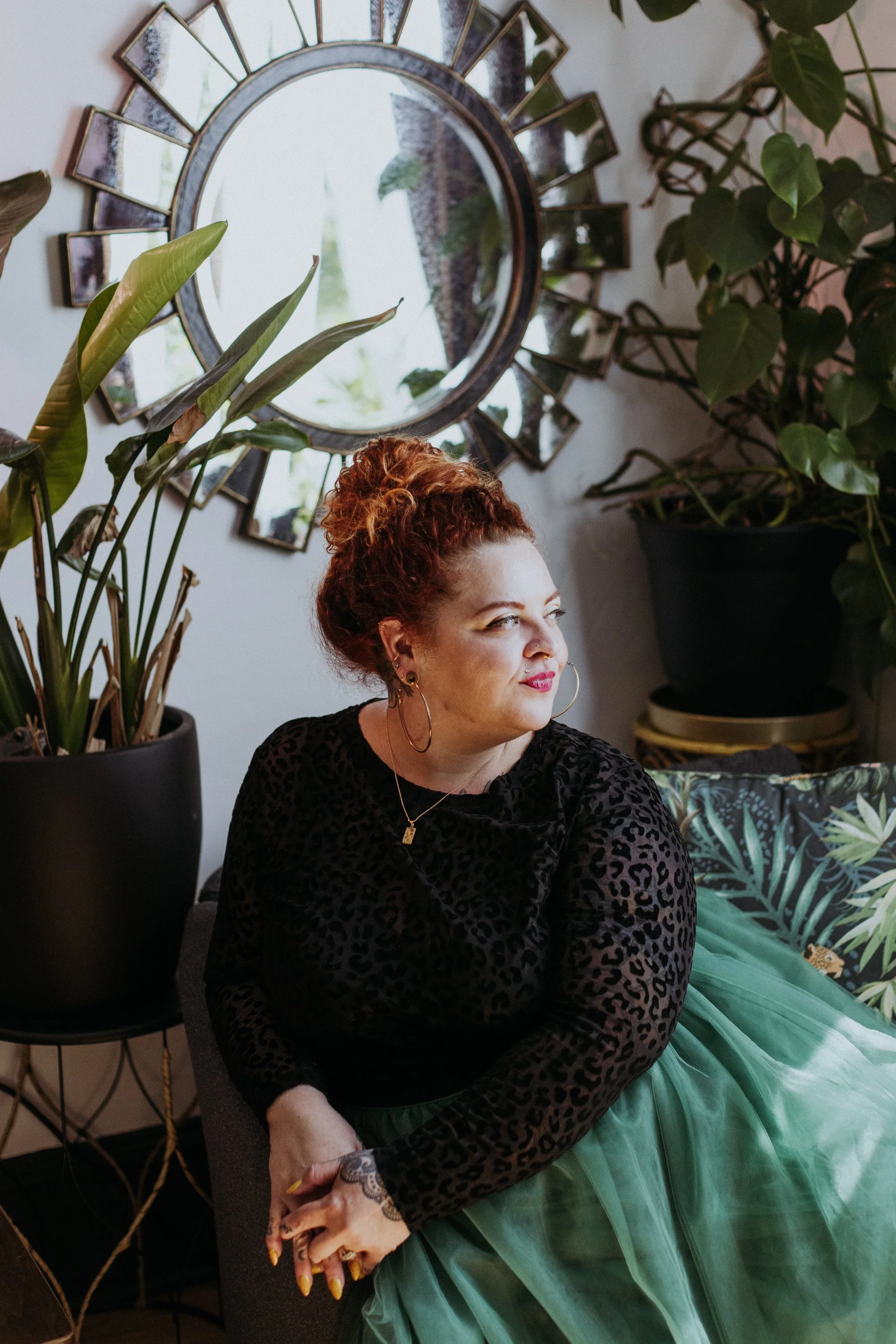 A woman with curly red hair, wearing a black leopard print top and a teal tulle skirt, is sitting in a room with potted plants and a decorative mirror on the wall. She has makeup, jewelry, and a tattoo on her hand.