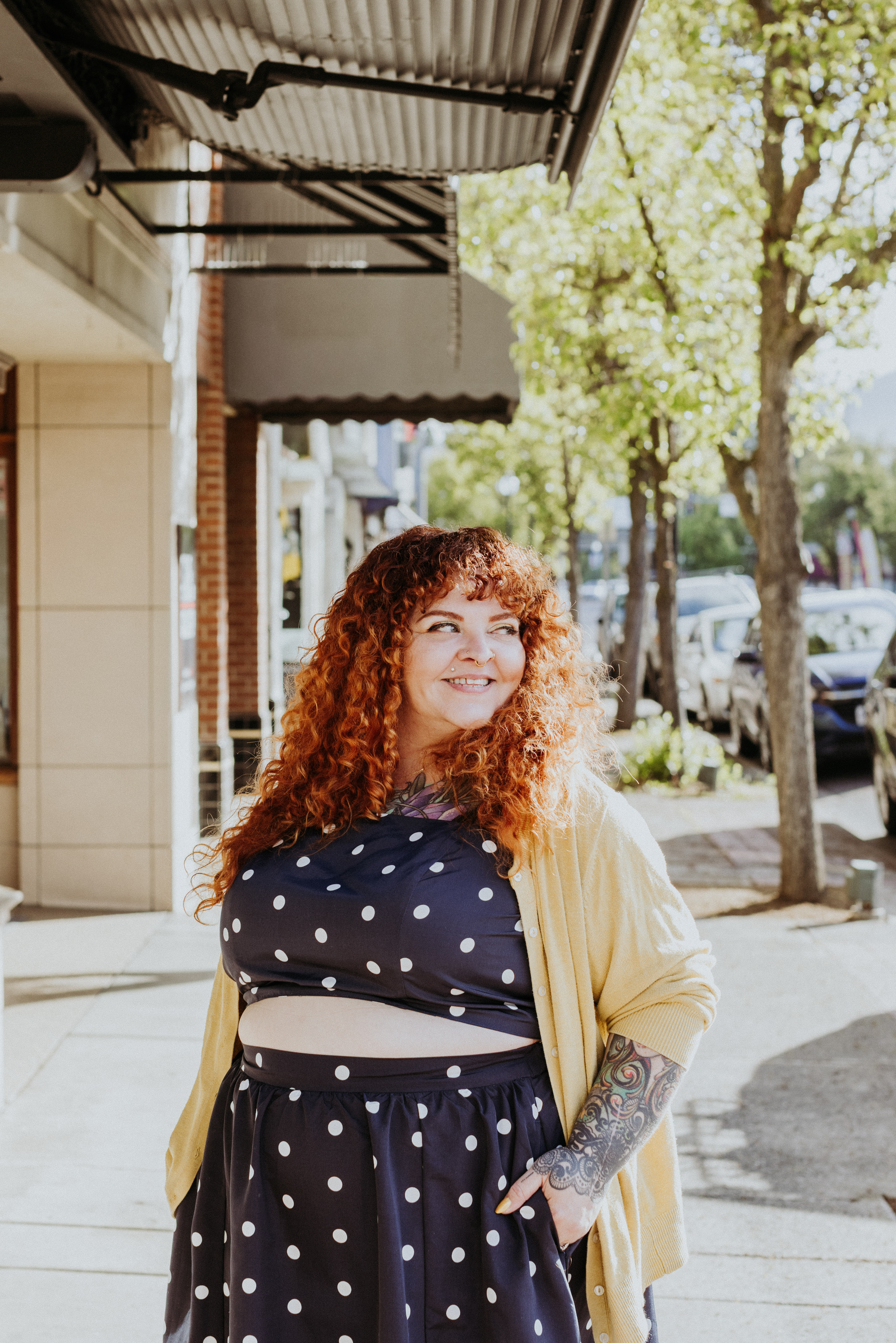 A woman with red curly hair and tattoos on her left arm is standing outside on a sunny day, smiling. She is wearing a navy blue dress with white polka dots and a yellow cardigan. There are trees, parked cars, and storefronts in the background.