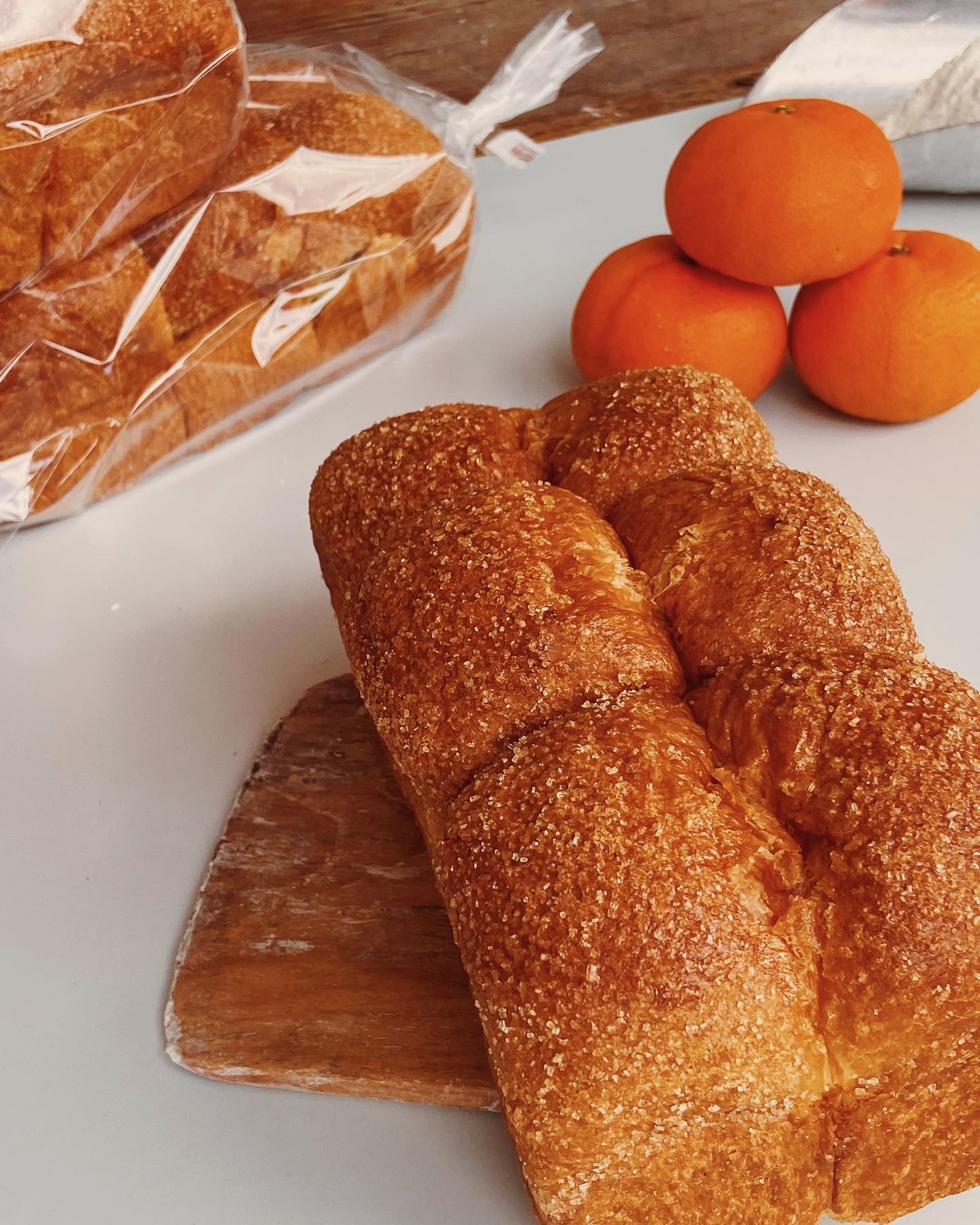 Sweet braided bread coated with sugar dusting on a white plate, with clementines and a loaf of bread in plastic packaging in the background.
