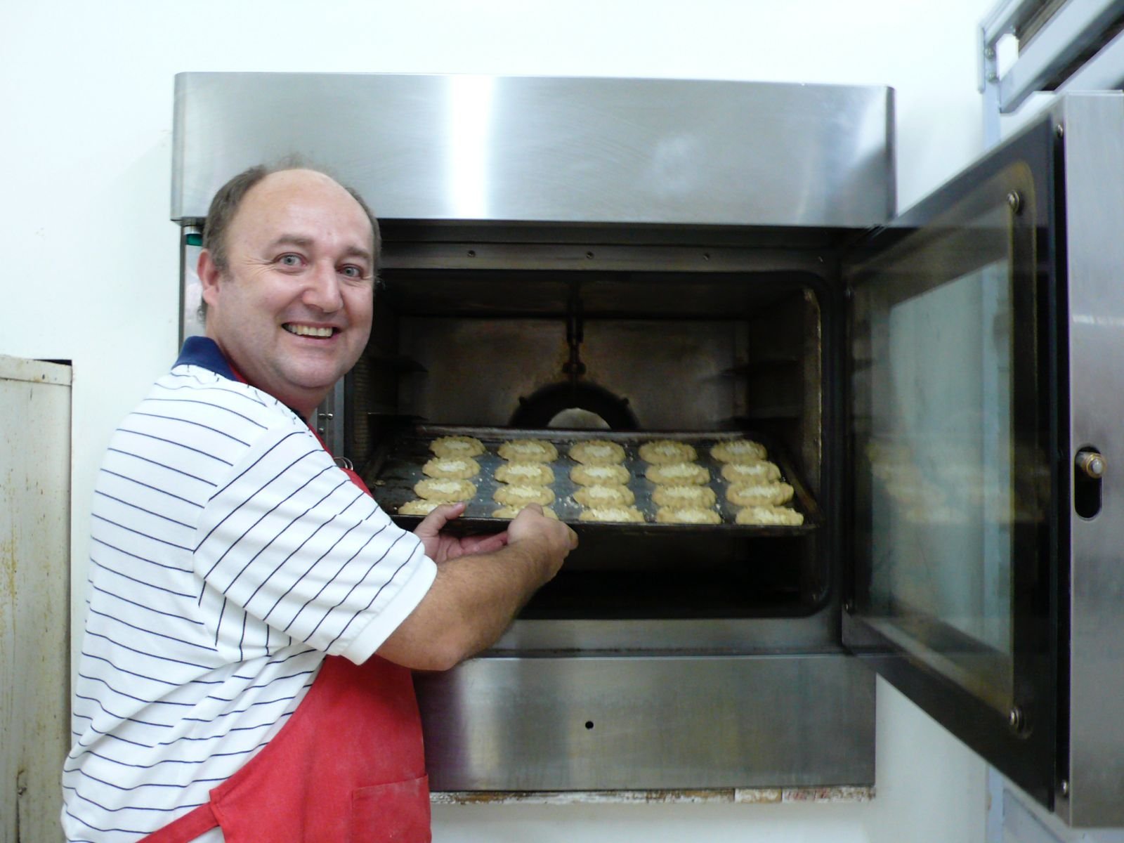 A man with a striped shirt and red apron placing a tray of raw food, possibly cookies or meat, into an industrial oven in a kitchen.