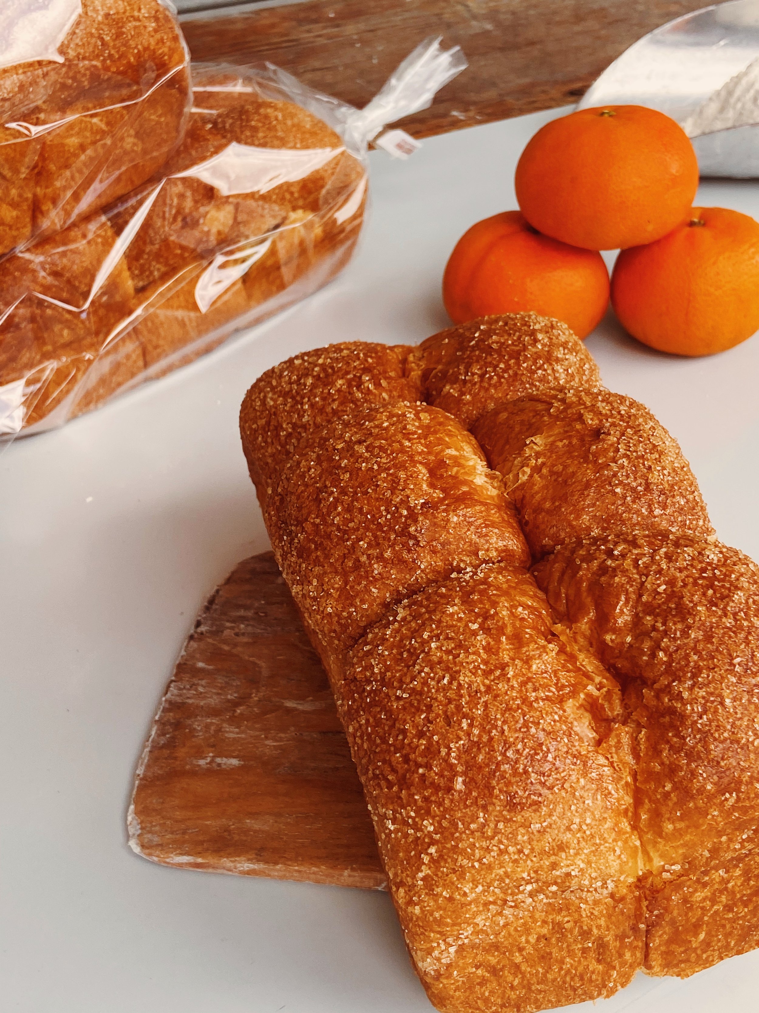 Fresh bread loaf on a wooden cutting board, with oranges and packaged bread in the background.