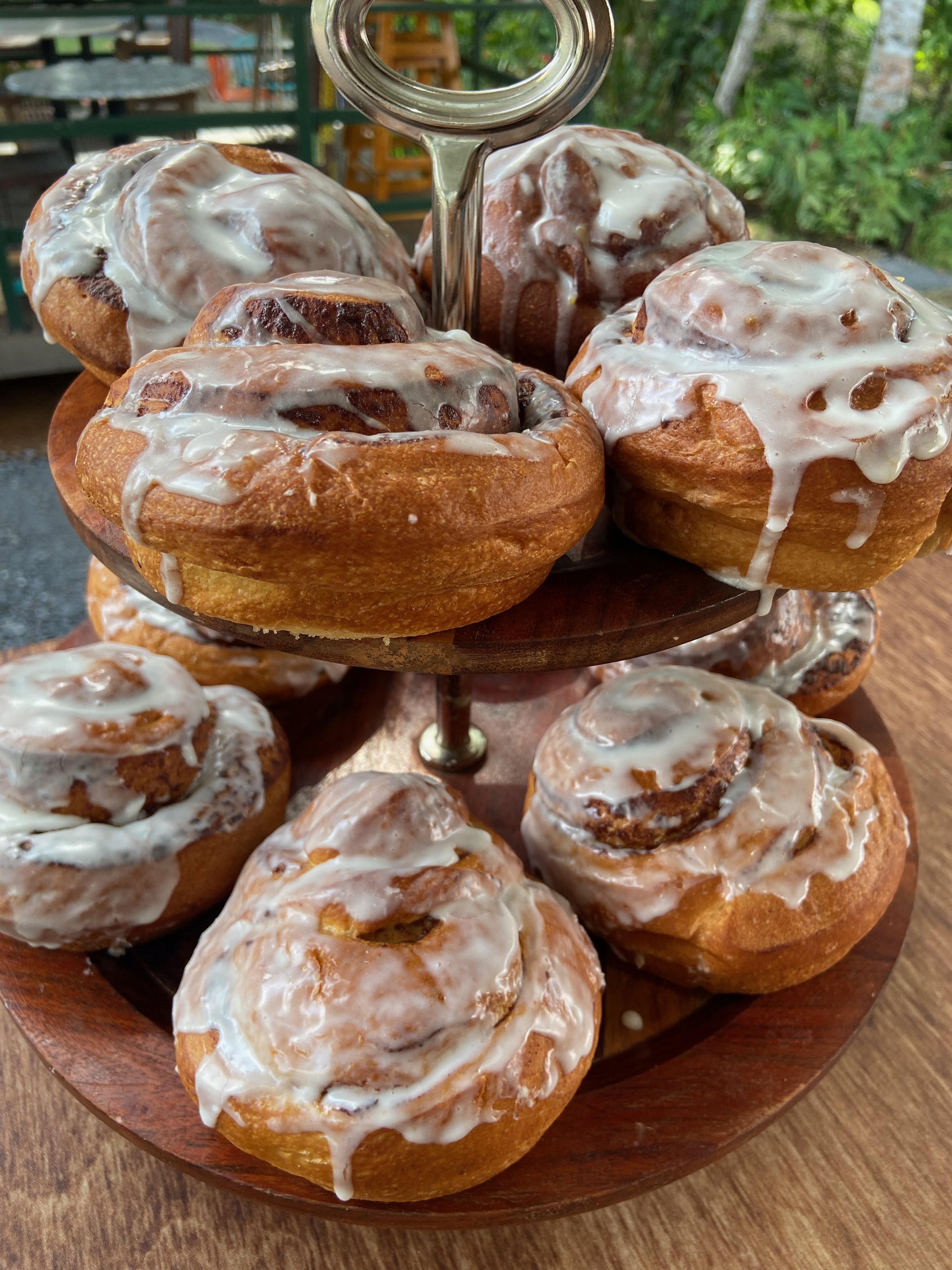 A wooden stand with cinnamon rolls topped with white icing on a wooden table outdoors.