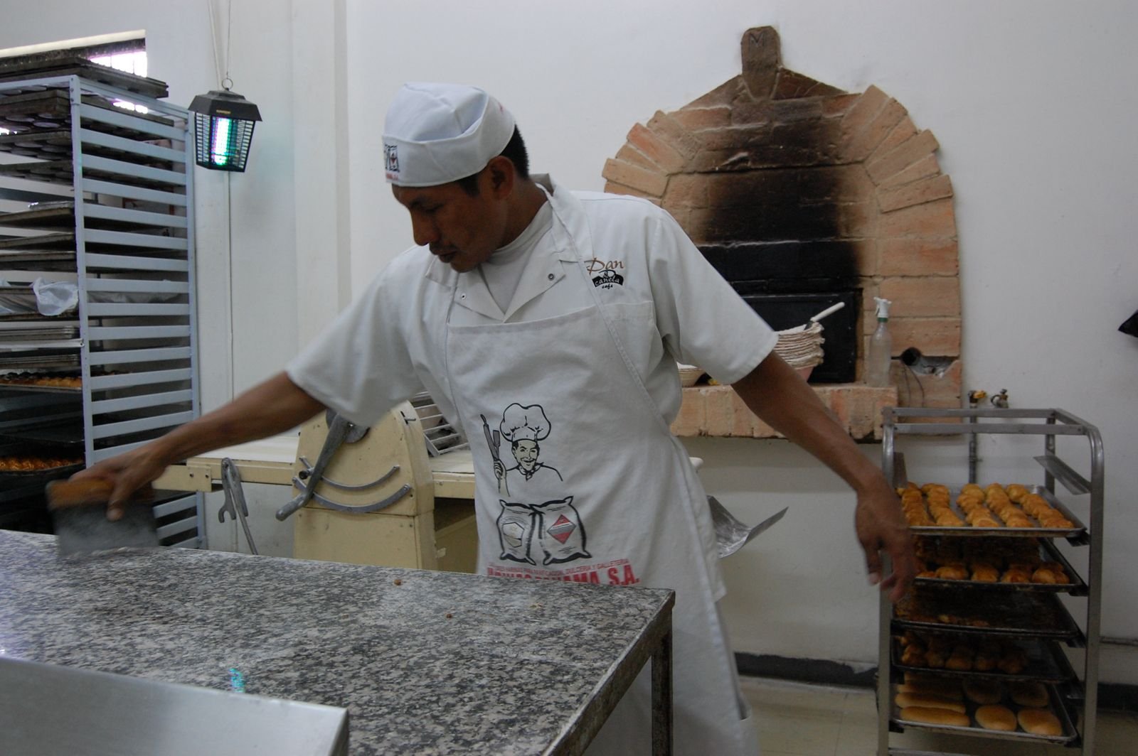 A baker in a white apron and hat working in a bakery kitchen with baked bread on a metal rack and a brick wood-fired oven in the background.