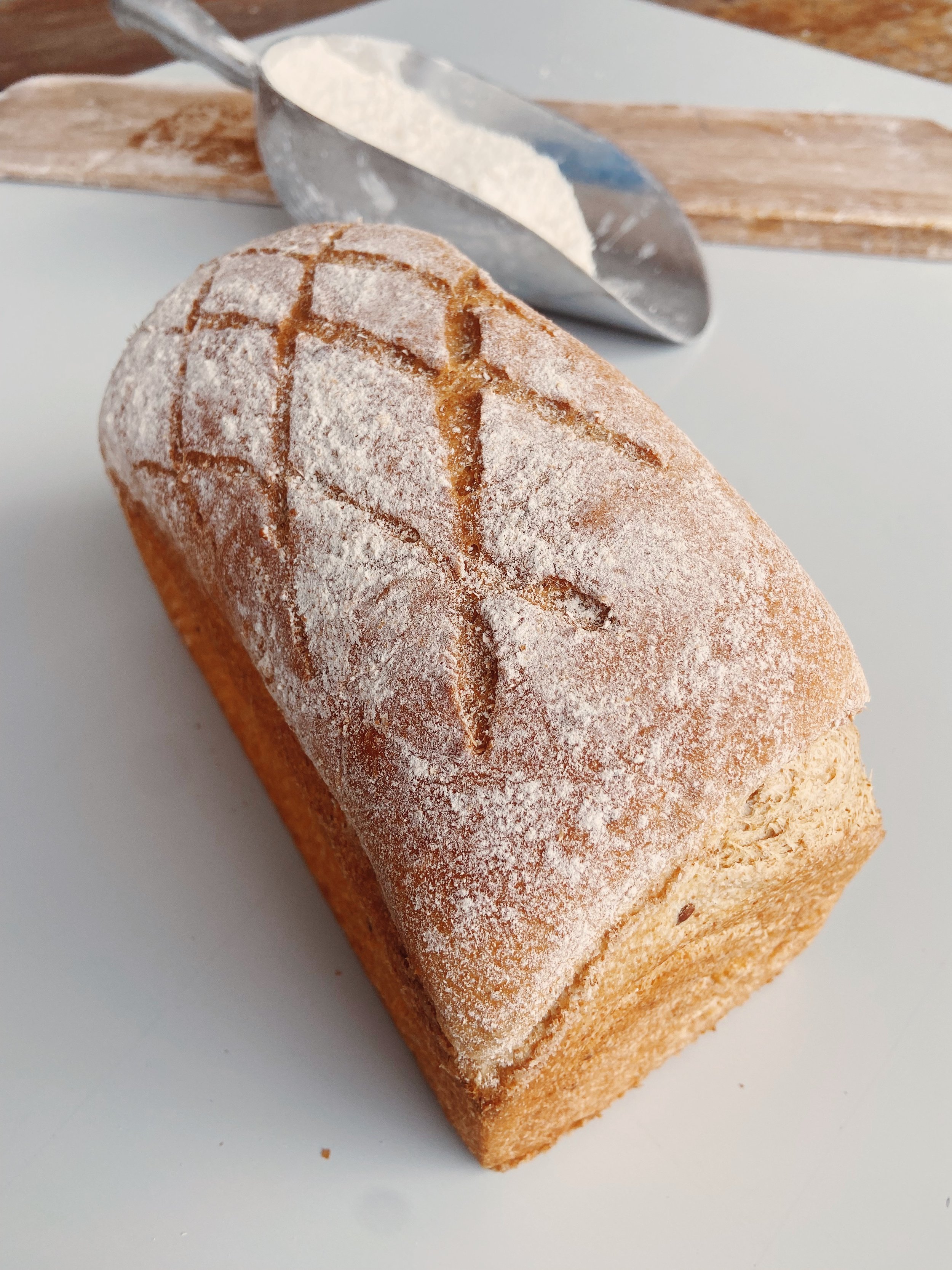 A freshly baked loaf of bread with a dusting of flour and a cross-shaped score on top, placed on a white surface with a bread scoring tool and flour in the background.