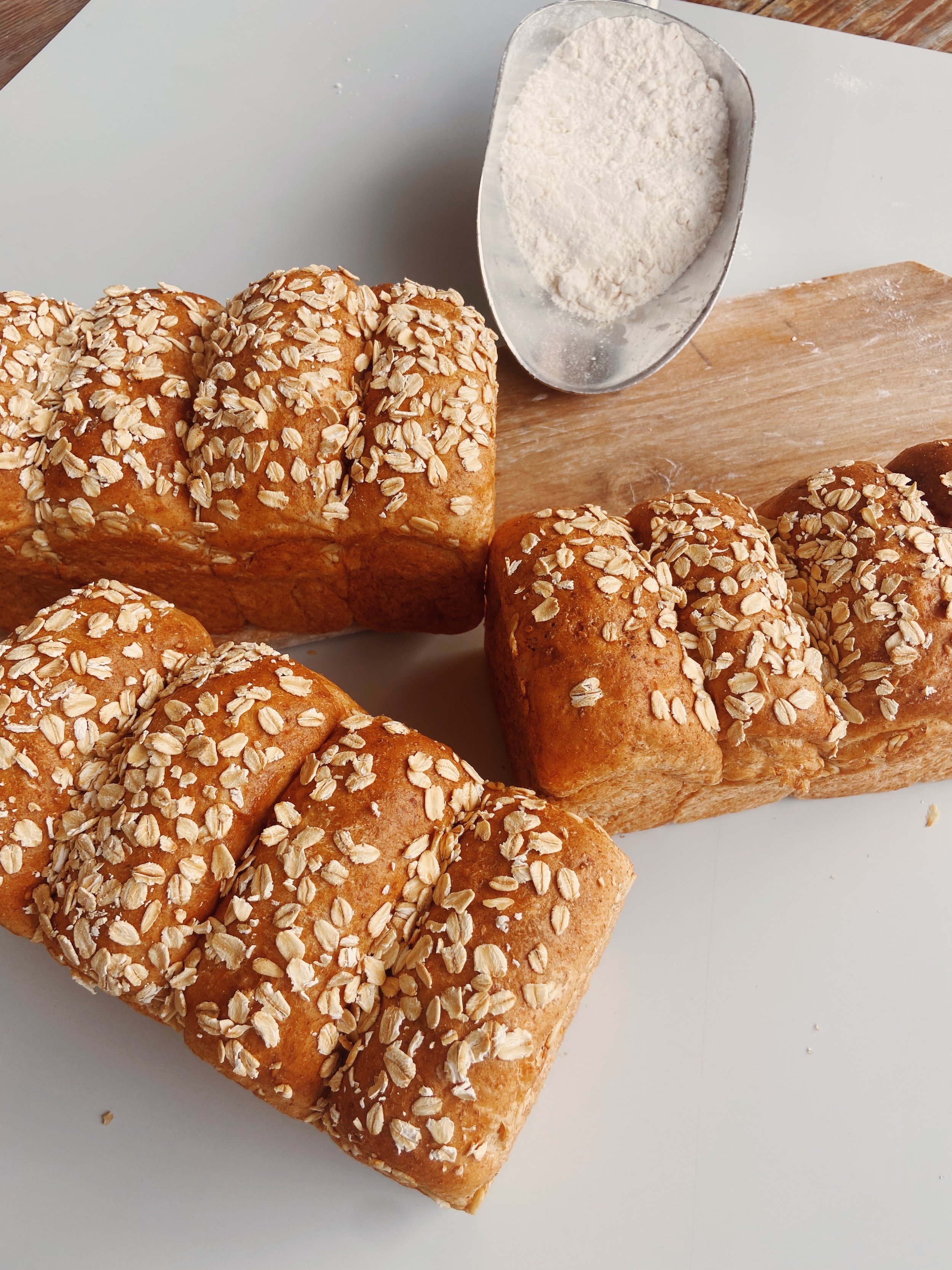 Three loaves of bread topped with oats, a metal bowl of flour, and a wooden cutting board on a white surface.