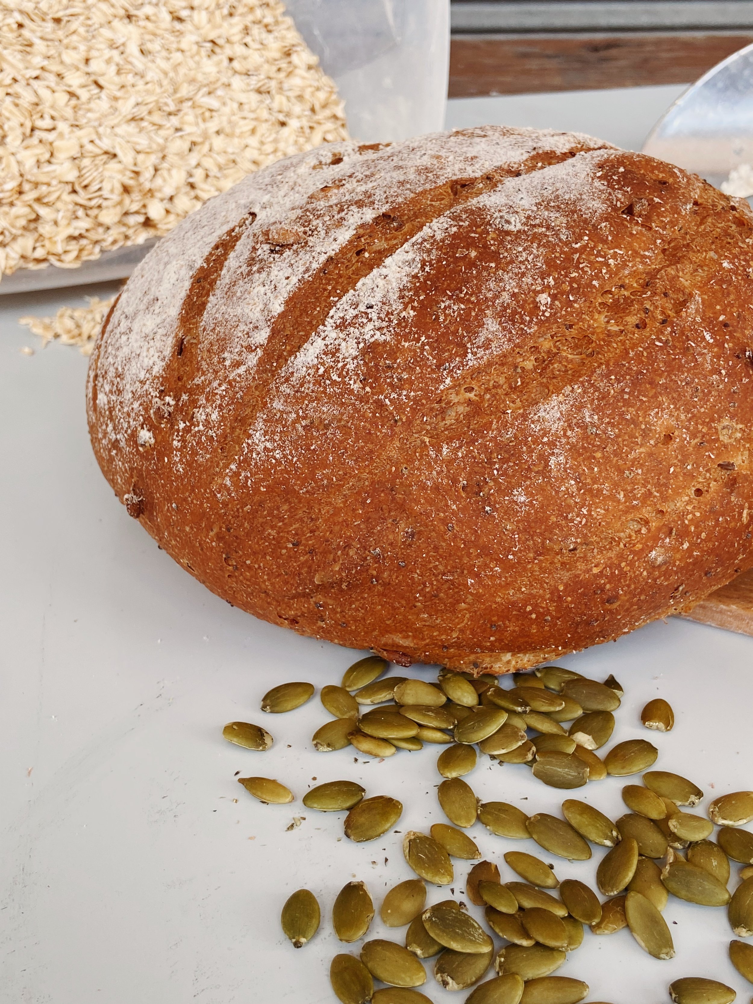A loaf of bread with a dusting of flour, surrounded by pumpkin seeds on a white surface, with grains in a container and a glass in the background.