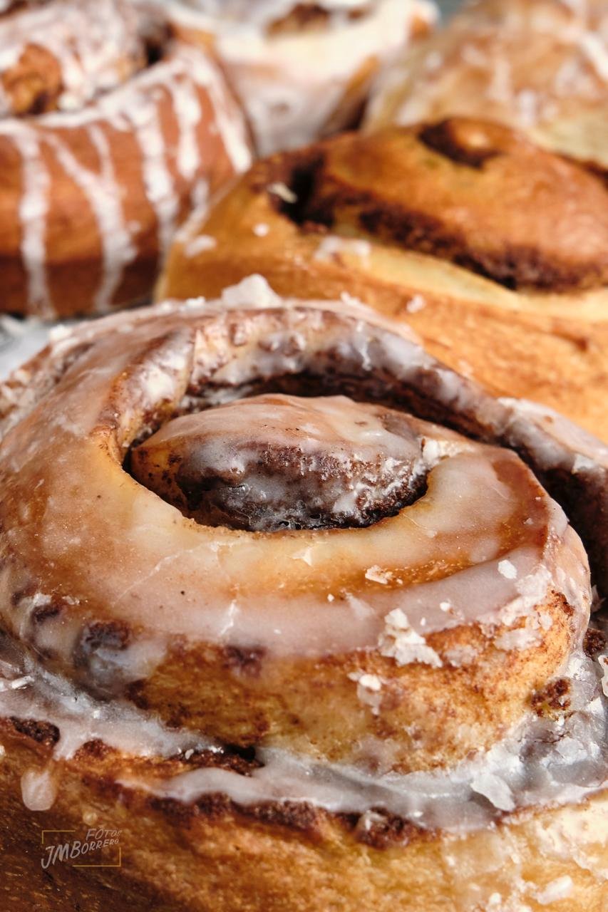 Close-up of a cinnamon roll with icing, topped with a chocolate chip, and other baked goods in the background