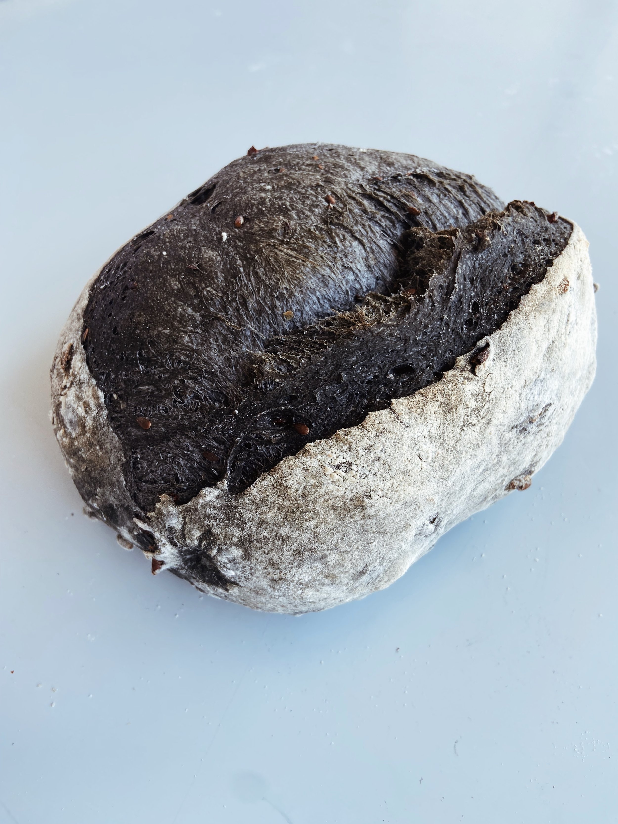 A round loaf of bread with a dark, glossy top and a lighter, flour-dusted bottom, sitting on a white surface.