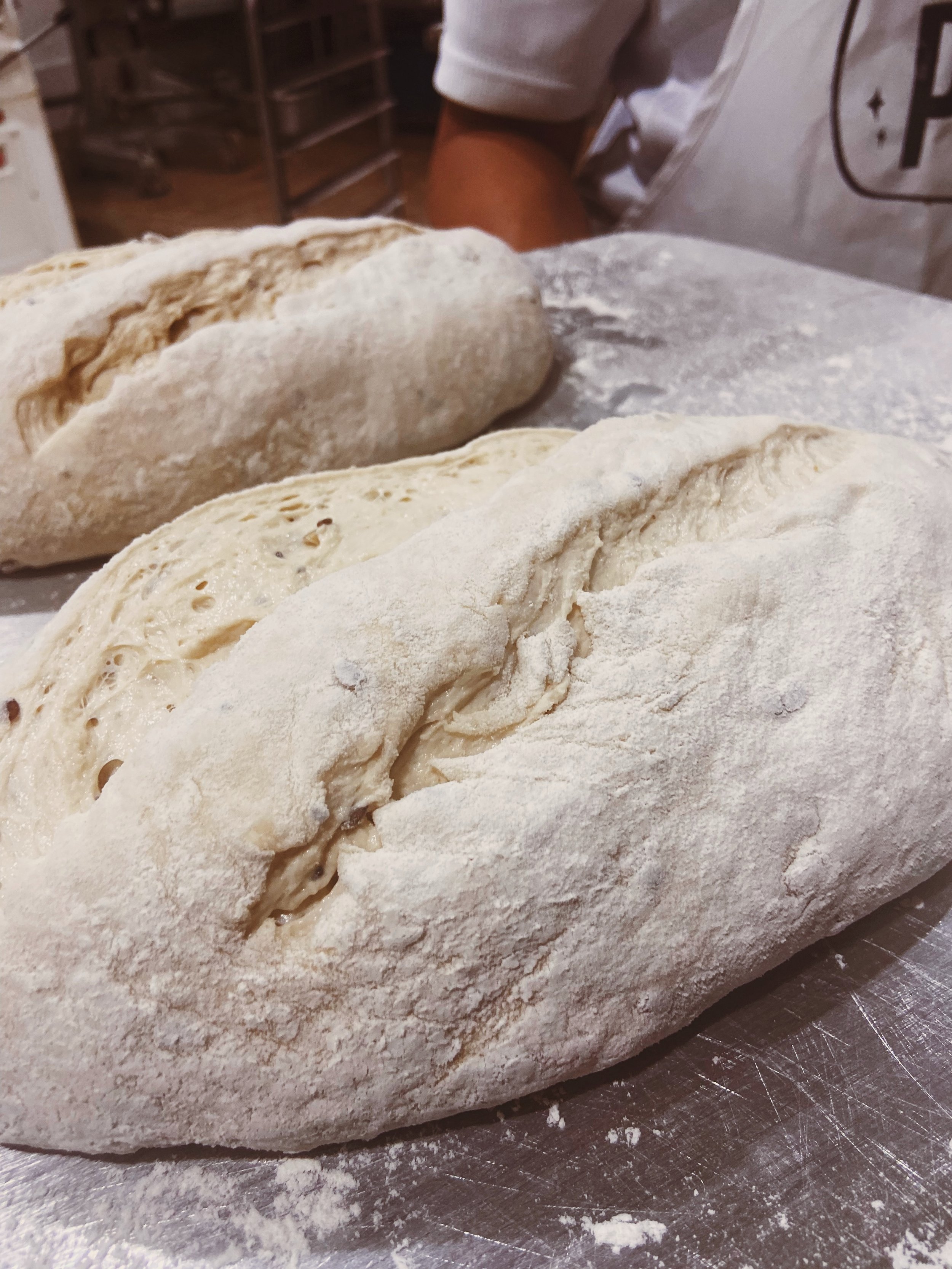 Two uncooked loaves of bread dough covered in flour on a baking sheet in a bakery setting.
